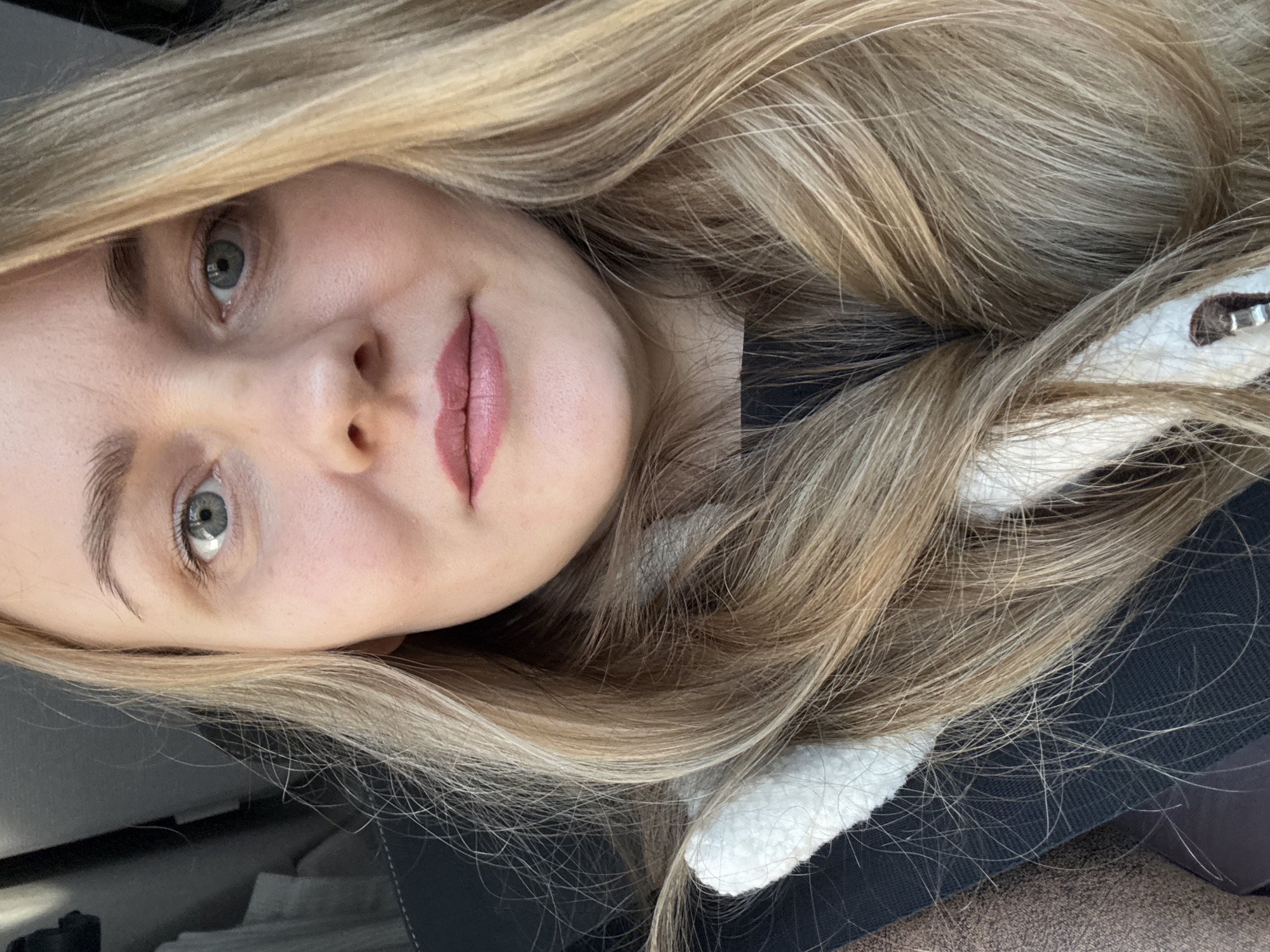 A close-up, eye-level portrait shows a light-skinned adult female inside a vehicle in Bolgury, Russia. She has long, wavy blonde hair with lighter highlights, parted on the left side and cascading over her shoulders and chest. Her eyes are blue-gray, and her light brown eyebrows are visible. Her lips are painted with a muted pink lipstick. Faint shadows are present beneath her eyes. She is wearing a dark-colored top, over which a cream or white fuzzy collar or trim is visible. A dark seatbelt strap crosses diagonally over her left shoulder and chest. A dark vehicle interior, including a dark seatback and what appears to be a headrest, is visible in the blurred background to the left. A textured brown fabric, possibly part of a coat or car seat, is visible on the far left edge of the frame. The subject is looking directly into the camera.