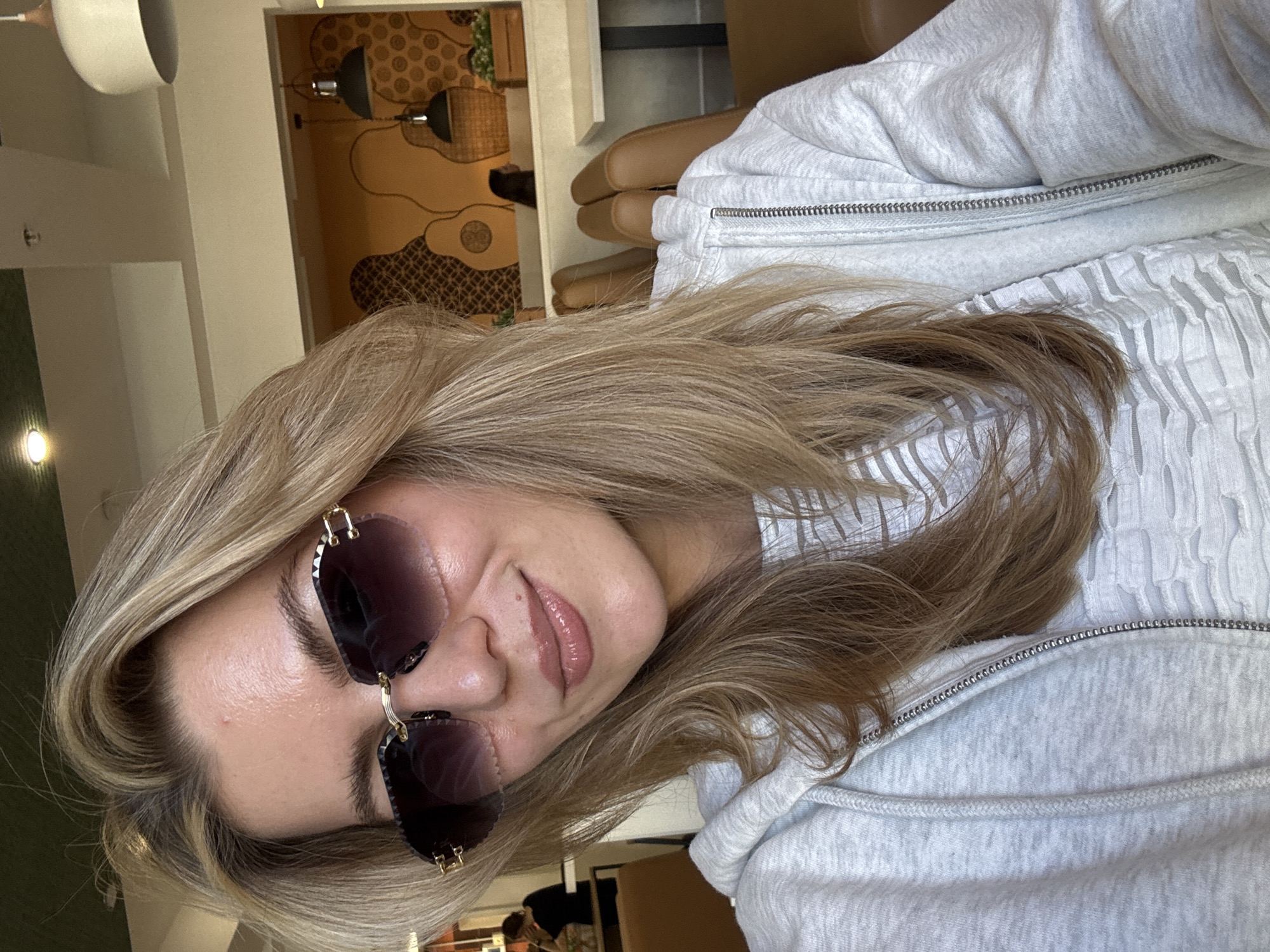 The image shows a person indoors, likely in a café or similar establishment. The person has blond hair, is wearing sunglasses, and is wearing a light gray zip-up sweater. The background includes a wall with patterned artwork and a view of the room, including furniture and lighting fixtures. The location in the city is unknown.