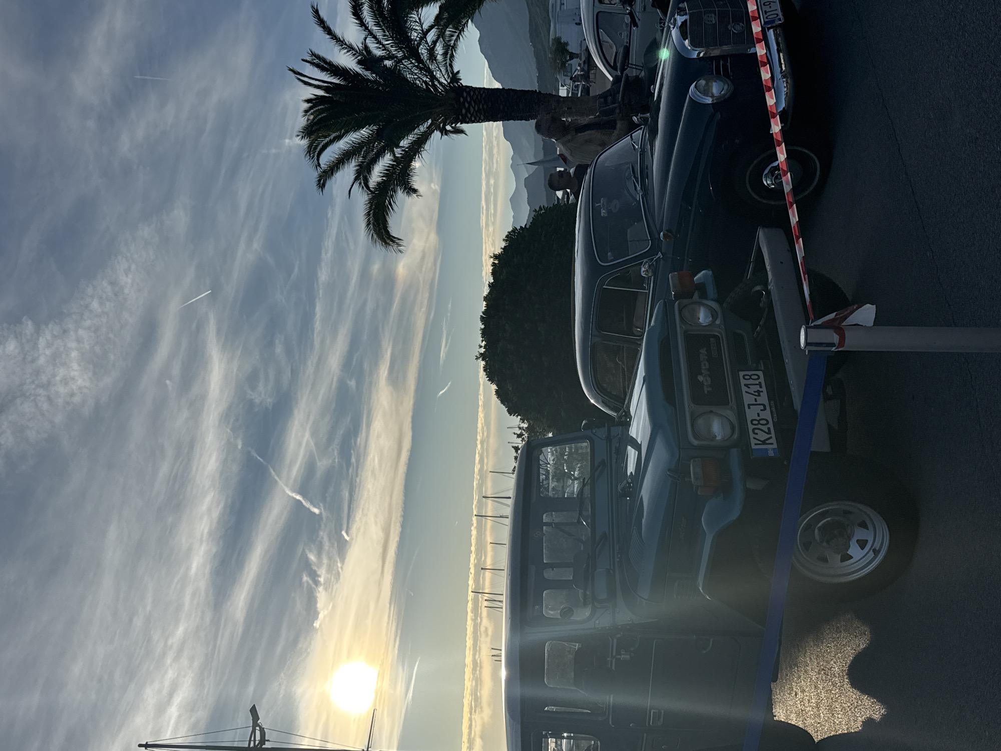 The scene depicts an outdoor classic car display at sunset, located by a marina in Bar, Montenegro. The sun, a bright orb with a prominent lens flare, is low on the left horizon, casting golden light and long shadows, consistent with late afternoon or early evening. The sky is largely clear blue with numerous wispy cirrus clouds and multiple aircraft contrails.
In the foreground, two classic cars are prominently featured and cordoned off by barrier tapes. On the left is a dark green Toyota Land Cruiser, likely an FJ40/BJ40 series, with a visible Montenegrin license plate "K28-J-418". A blue barrier tape runs horizontally across the lower part of the image, in front of this vehicle. To the right, slightly behind the Toyota, is a dark-colored Mercedes-Benz sedan, identifiable by its grille and headlights, possibly a "Fintail" model. Its license plate is partially visible, reading "OT-9". This Mercedes is cordoned off by a red and white striped barrier tape, attached to a white and red striped pole in the lower foreground.
Several individuals are visible in the mid-ground. One person in a light-colored jacket stands near the front passenger side of the Mercedes-Benz, appearing to observe the vehicle or converse with another person partly obscured behind them. Other indistinct figures are present further in the background.
The environment includes a tall palm tree with a robust trunk on the right side of the frame. To the left of the palm tree is a dense green bush or tree. In the middle distance, numerous masts of sailboats are visible, indicating a marina or harbor. A range of mountains, appearing hazy in the distance, forms the backdrop. The ground surface appears to be paved asphalt, reflecting the low sunlight in some areas. The "BR" insignia on the Toyota's license plate confirms the location as Bar, Montenegro.