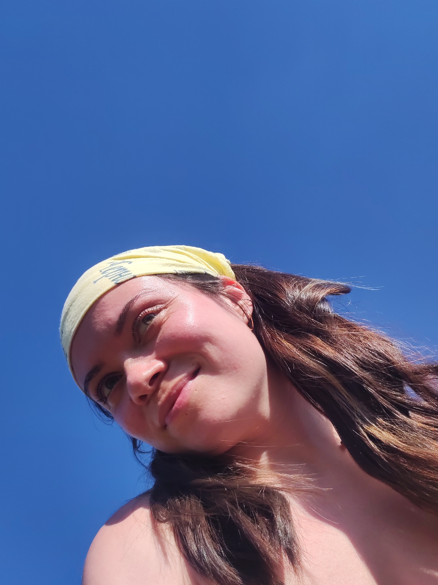 A low-angle shot of a person with fair skin, brown hair, and green eyes against a clear, bright blue sky. The person is wearing a yellow headband with light-colored lettering on it. The person is smiling slightly. The location cannot be ascertained as Фоки, Russia without further context.