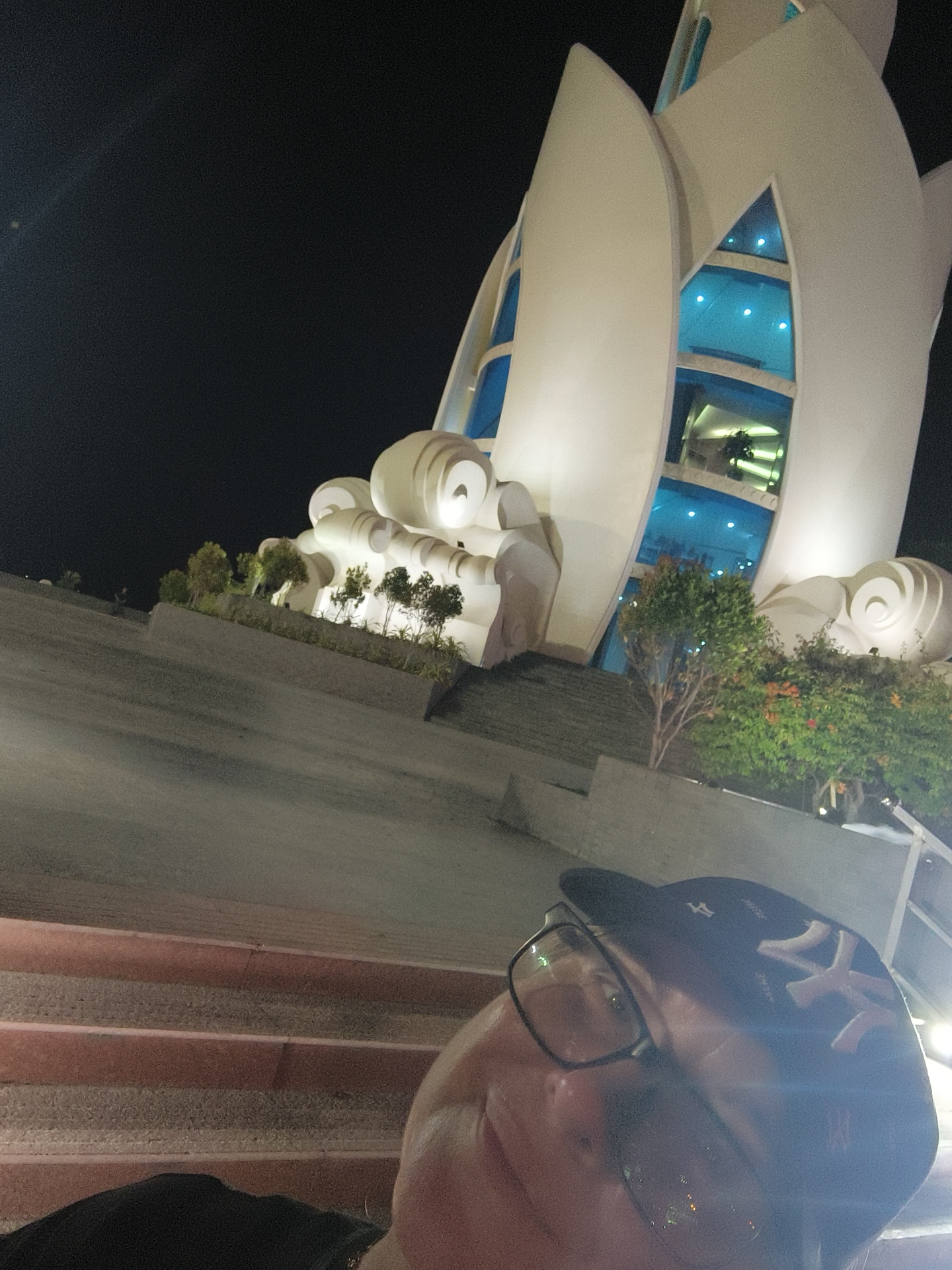 The image shows a person wearing glasses and a New York Yankees baseball cap, looking up at the entrance of a modern building with a unique, petal-like architectural design at night. The building appears to be illuminated, with blue windows and internal lights visible. The scene is set in Nha Trang, Vietnam, likely at the Vinpearl Land Amusement Park or a similar resort area, given the architectural style and the presence of landscaping. The person is in the foreground, with stairs leading up towards the building. The lighting suggests it's evening or night.