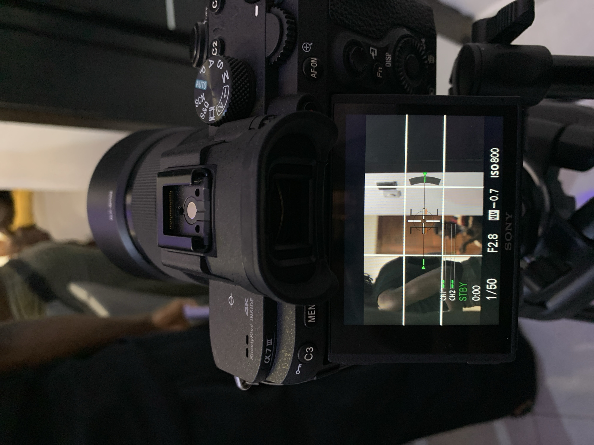 A black Sony Alpha 7 III mirrorless camera, equipped with an attached Sony FE 24-70mm GM lens, is depicted from a slightly elevated rear perspective. The camera's mode dial is set to "AUTO," and "4K SteadyShot INSIDE" is visible on the top plate. The articulating LCD screen is active, displaying a live view grid overlay with a person's upper torso and arm in a dark garment positioned on the left side of the frame, against an interior background featuring a light-colored wall or doorway. Visible camera settings on the LCD include: STBY status, 0:00 timer, 1/50 shutter speed, F2.8 aperture, -0.7 exposure compensation, and ISO 800, along with green audio level indicators for CH1 and CH2. A central crosshair focus target is displayed. The camera is mounted on a black tripod, partially visible at the bottom right. Blurred human figures are present in the immediate background behind the camera. The scene is located in the city of Igboefon, Nigeria.