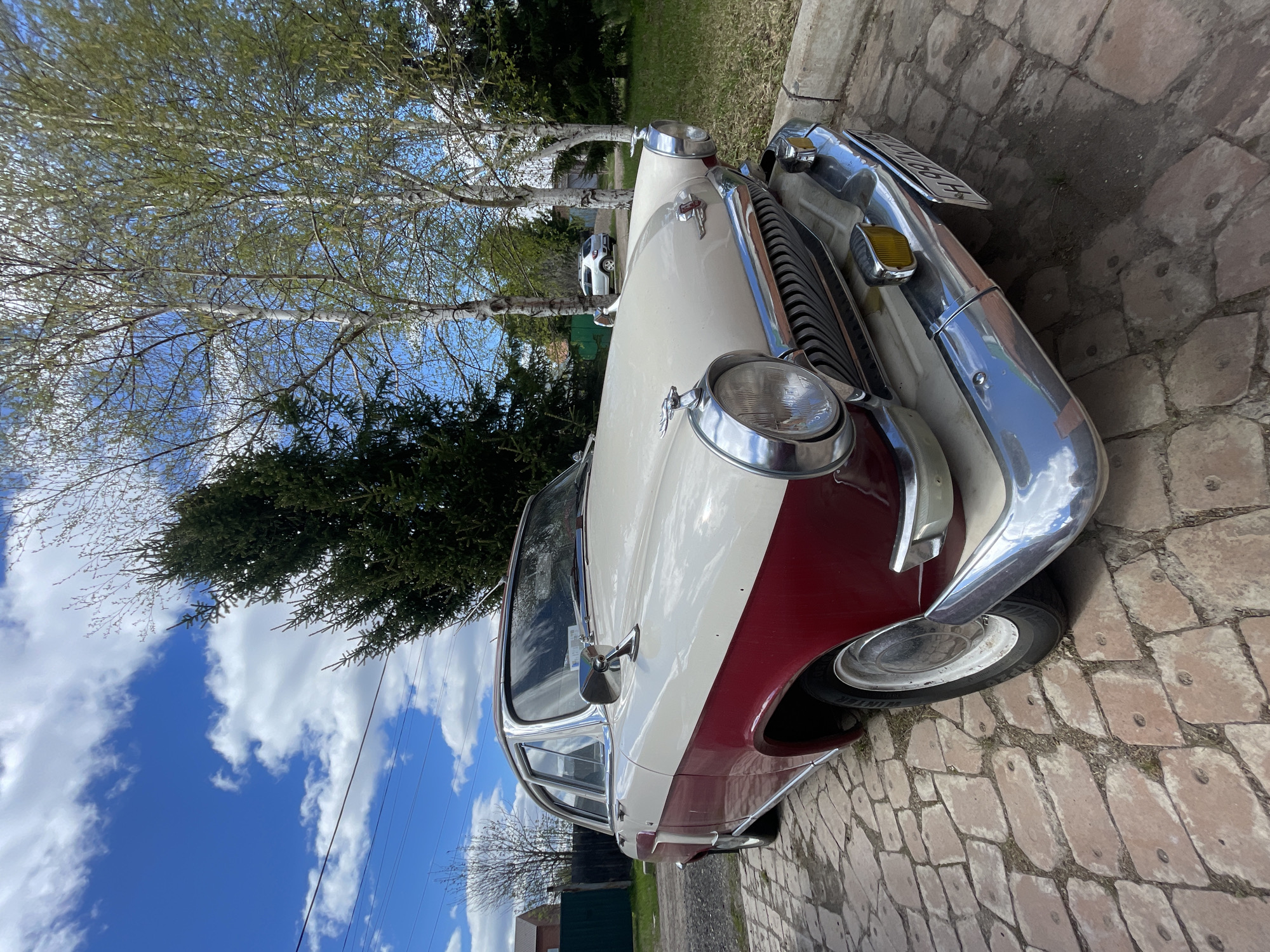 A low-angle, full outdoor shot shows a vintage two-tone (cream/red) automobile, possibly parked. The right front end of the vehicle is in the immediate foreground. The car is parked on a cobblestone surface. The sky, partially obscured by sparse trees, is bright blue with large, fluffy white clouds. A portion of the city Минино, Russia, is visible in the midground. The lighting is bright and even.