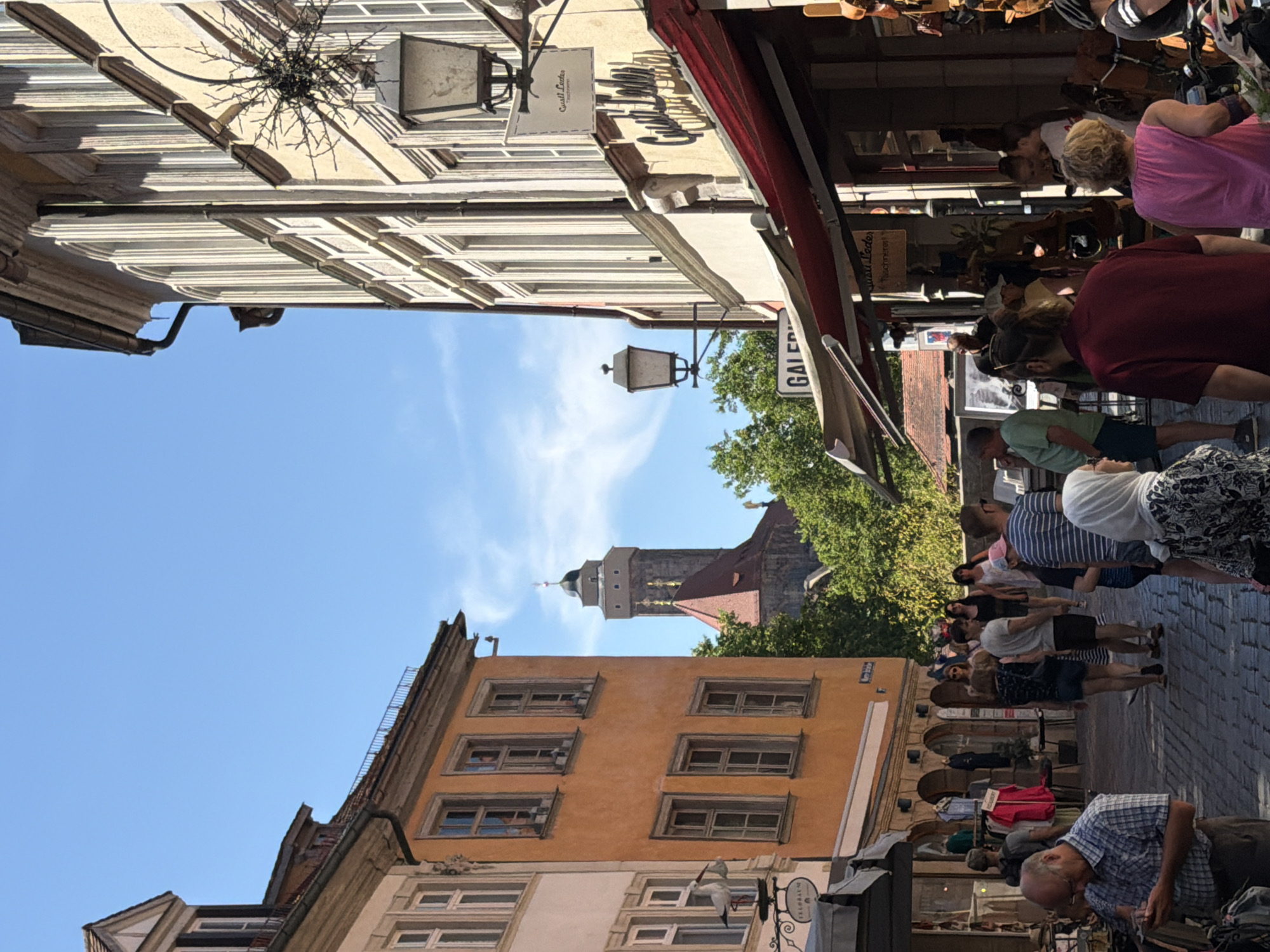 The image captures a vibrant street scene in Bamberg, Germany, likely during a sunny day. The architecture features traditional European buildings with colorful facades, particularly an orange building on the left adorned with windows and a whimsical stork figurine carrying a baby. On the right, a tall, cream-colored building with multiple arched windows and decorative elements looms. A street lamp with a star-shaped ornament is visible attached to this building. In the background, a prominent tower with a green spire, likely part of a historic church or cathedral, rises above lush green trees.
The street itself is paved with cobblestones and bustling with people. Shoppers browse stalls, some displaying clothing and other goods under awnings. Several individuals are walking along the street, engaged in conversation or looking at their phones. A man in a plaid shirt examines his phone in the foreground. A woman in a pink top and a woman in a white hijab are also visible among the crowd. The atmosphere appears lively and typical of a European market street.