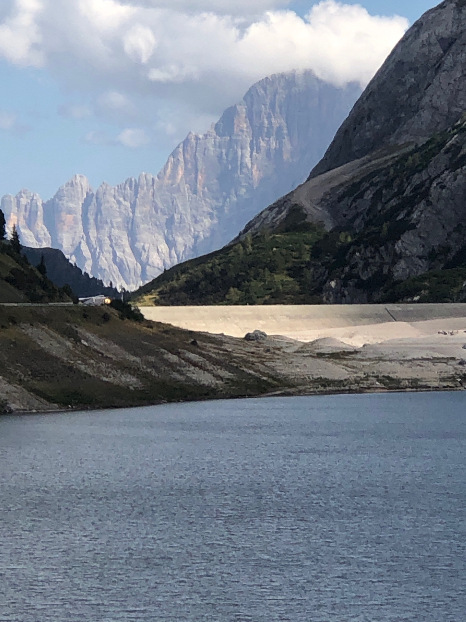 The image depicts a serene mountainous landscape with a body of water in the foreground. The water, a muted blue, exhibits gentle ripples, suggesting a calm environment. Behind the water, a large, light-colored dam stretches horizontally across the mid-ground, its smooth, concrete surface reflecting the soft daylight. The dam is situated at the base of a steep, rocky mountain slope that rises dramatically into the background. This slope is partially covered with sparse green vegetation, particularly on the left side of the frame.
Further in the distance, the majestic peaks of the Dolomites mountain range dominate the scene. These mountains are characterized by their rugged, stratified rock formations, with sharp ridges and vertical cliffs. The peaks are bathed in a soft, diffused light, and some are partially obscured by wispy white clouds. A small, light-colored structure, possibly a building, is visible on the hillside to the left, near a road that winds along the contours of the terrain.
The sky above is a pale blue, adorned with scattered, fluffy white clouds that add a sense of depth and atmosphere to the expansive vista. The overall impression is one of natural grandeur and tranquility, with the man-made structure of the dam subtly integrated into the breathtaking natural setting. The location is Canazei, Italy, a well-known tourist destination nestled within the Dolomites, celebrated for its stunning alpine scenery and outdoor recreational opportunities.