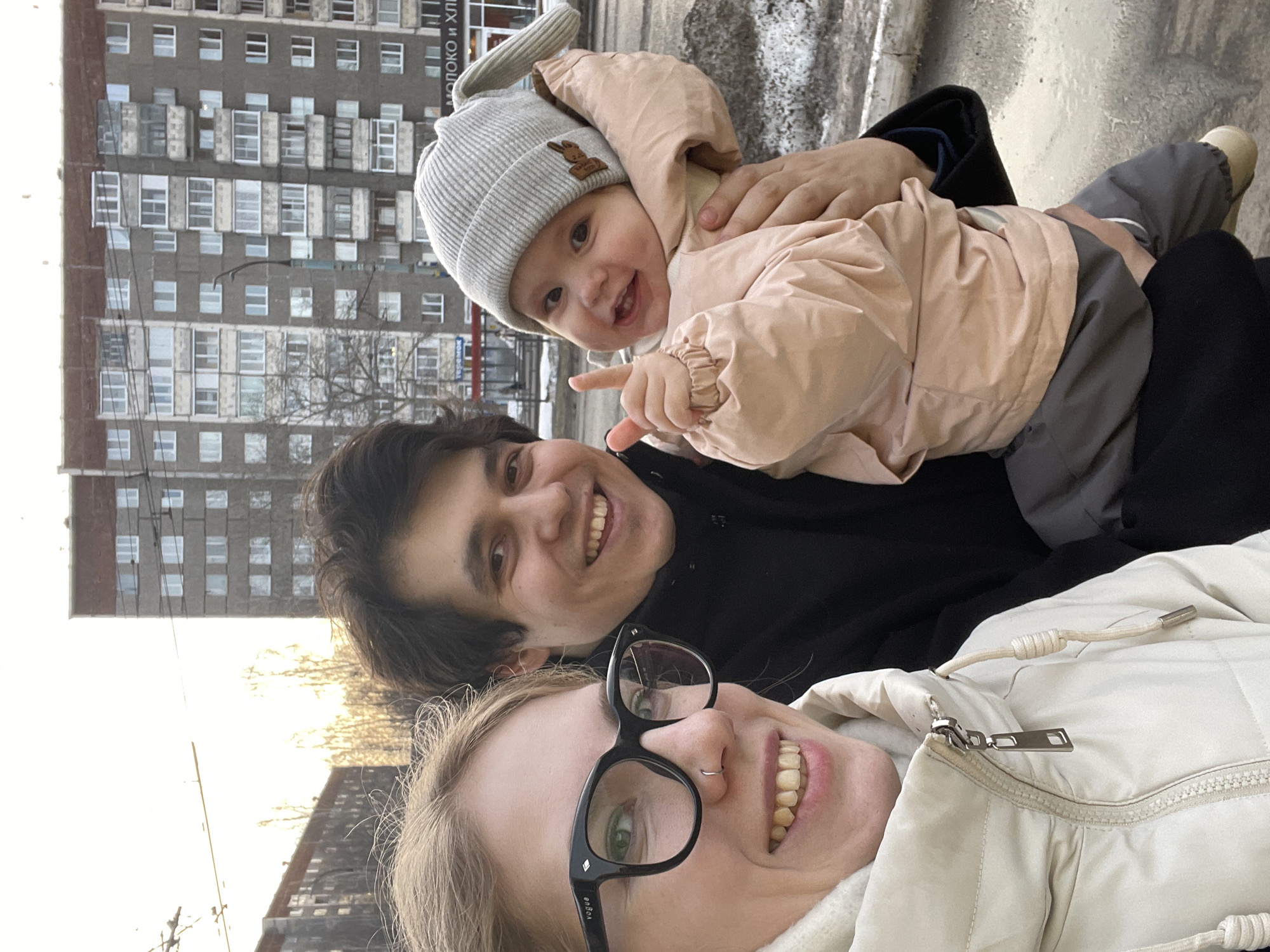 A selfie features three individuals—two adults and one infant—outdoors in Izhevsk, Russia. A blonde female adult, positioned on the left, wears black-framed glasses (brand "Vogue" visible) and a nose ring, dressed in an off-white padded winter jacket, smiling directly into the camera. To her right, a male adult with dark hair, wearing a dark coat, also smiles at the camera while cradling an infant. The infant, wearing a light grey ribbed knit hat with ear-like protrusions and a light pink padded jacket over dark grey pants, smiles broadly, looking at the camera, with its right index finger raised. The background consists of grey multi-story residential apartment buildings, characteristic of Soviet-era architecture, one displaying enclosed balconies. Patches of snow and slush are visible on the ground. Overhead trolleybus wires cross the bright, possibly late-afternoon, sky. A partially visible store sign on a background building reads "МОЛОКО И ХЛЕБ" (MILK AND BREAD) in Cyrillic.