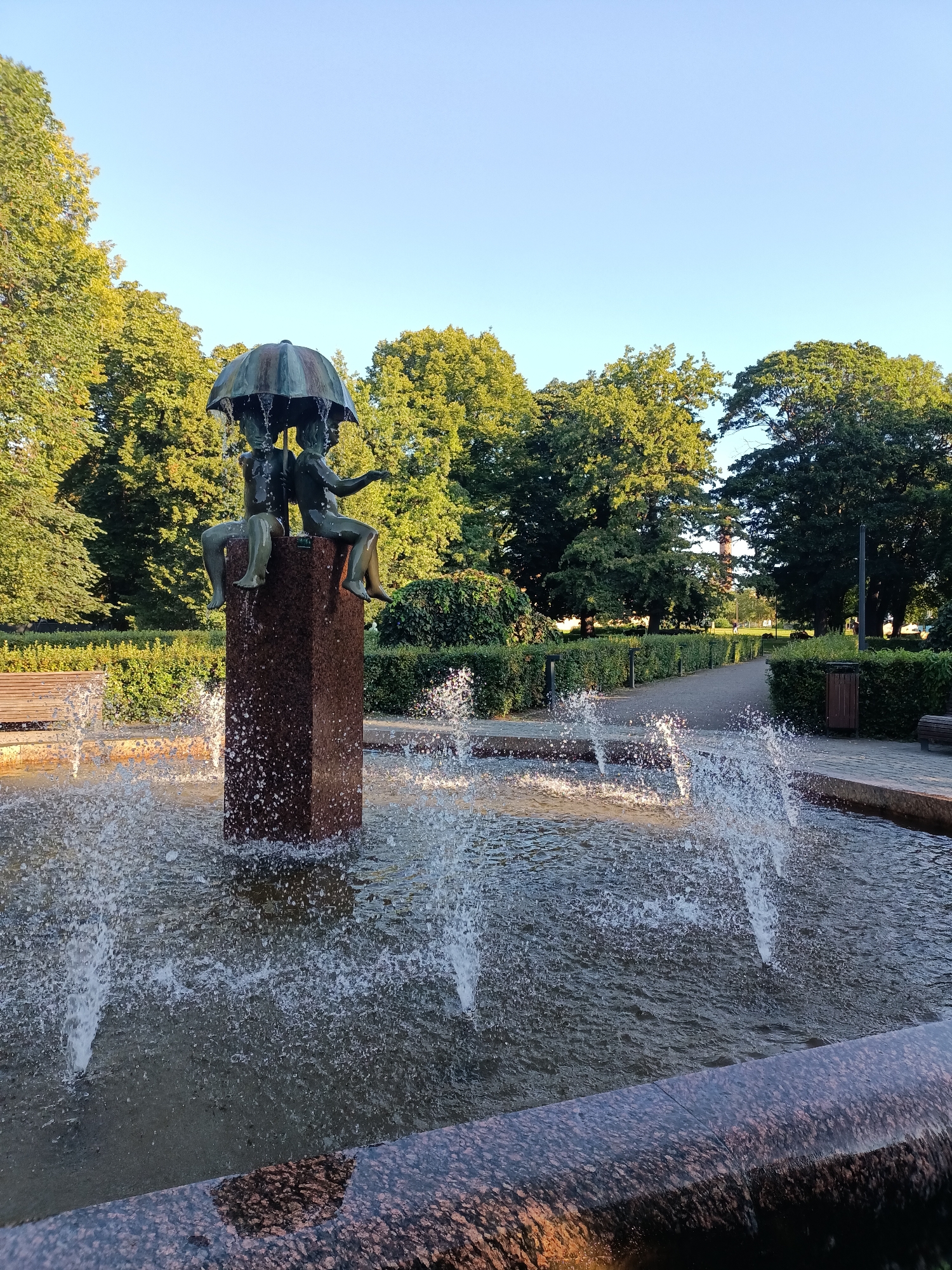 The image shows a fountain in a park in Tallinn, Estonia. The fountain features a bronze sculpture of two children sitting under an umbrella, with water pouring from the umbrella. The sculpture is placed on a red granite pedestal in the center of the fountain. The park is lush with green trees and manicured hedges. A paved pathway runs through the park, with benches and trash cans visible along the sides. The sky is clear and blue, indicating a sunny day. The water from the fountain is actively spraying, creating a dynamic effect. The overall scene depicts a serene and pleasant park environment.