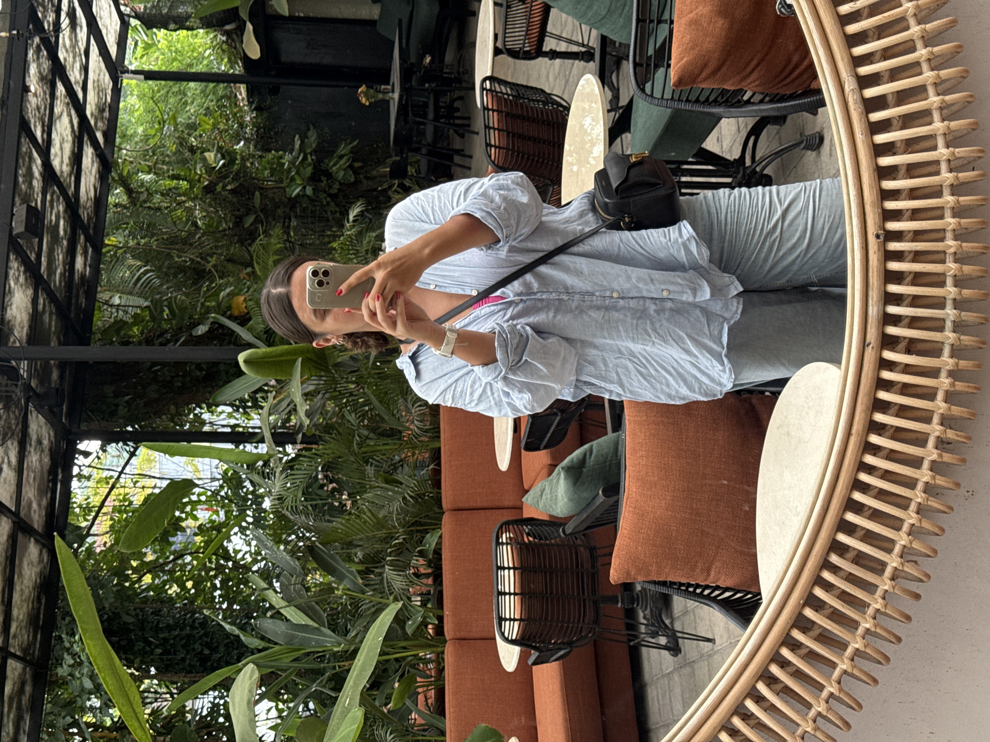 The image shows a young woman in a light blue, button-down shirt and jeans taking a selfie in a circular, rattan-framed mirror. She is holding a smartphone and looking at the screen. The scene is set in a lush, tropical location, likely a cafe or restaurant, given the chairs and tables visible in the background and mirror reflection. The reflection also reveals brown and green cushions on the chairs. Abundant greenery surrounds the seating area. The location is identifiable as the Ubud District, Indonesia, due to the distinctive tropical plants, the architecture, and style of furnishings.