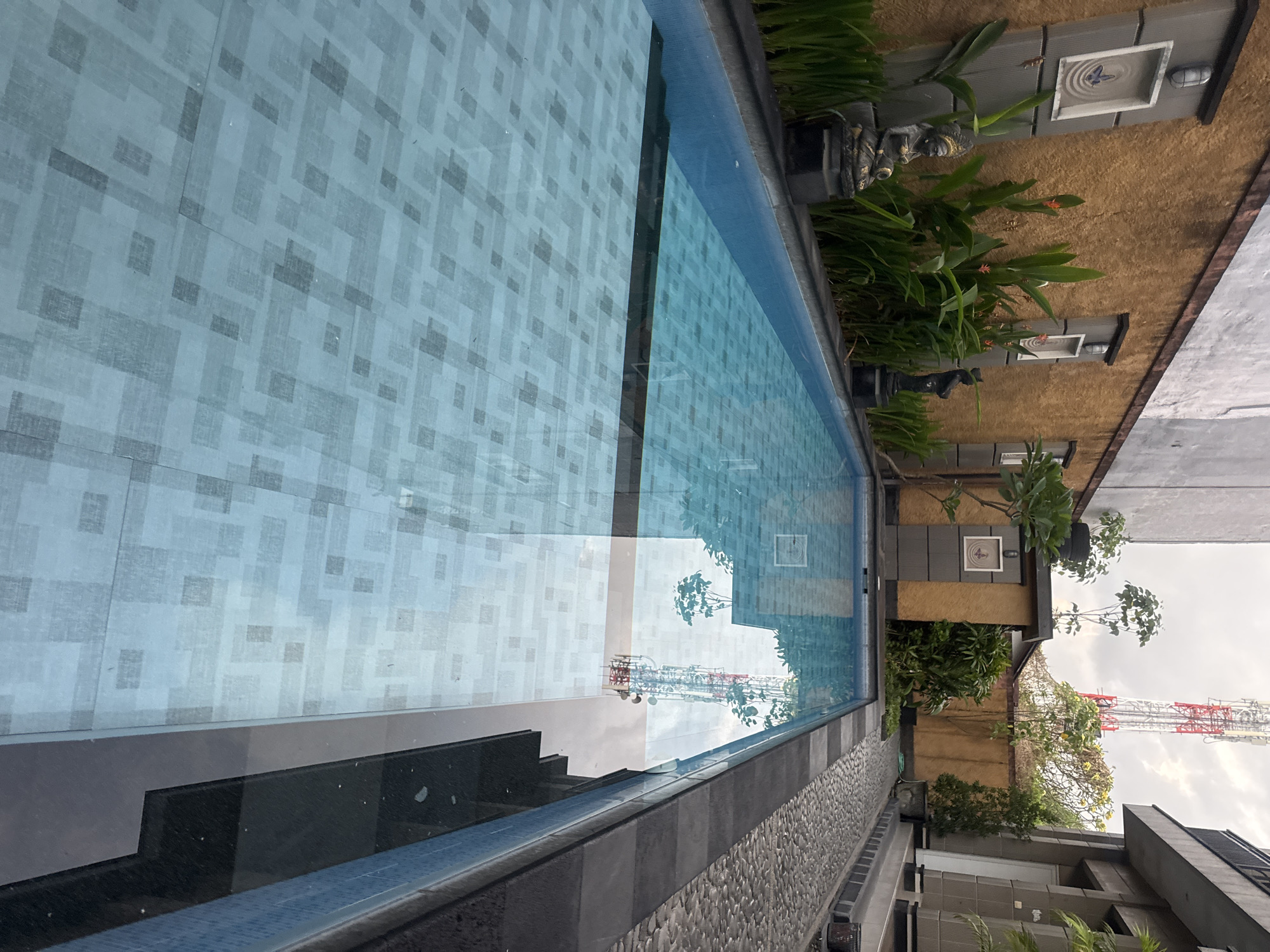The image depicts a swimming pool area with a tiled bottom, surrounded by a paved walkway and a textured yellow wall with decorative elements. To the left, two statues stand near the pool's edge, accompanied by lush green plants. In the background, a tall telecommunications tower is visible, along with buildings and trees. The scene appears to be from Kuta, Indonesia, given the architectural style and vegetation. The water in the pool is clear, with reflections of the surroundings visible on its surface.