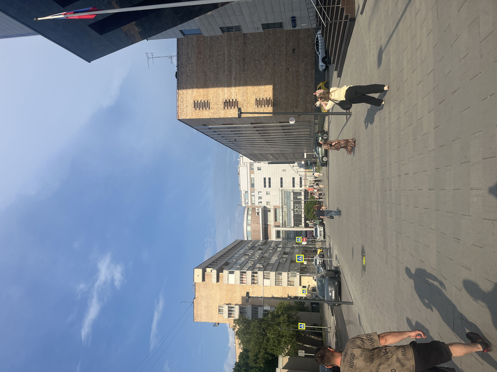 The image captures a street scene in Moscow, Russia, likely near a modern commercial or residential complex. In the foreground, a man wearing a tan t-shirt with a repeating "HUGO" pattern and black shorts walks away from the camera, casting a long shadow. To his right, a woman in a long, patterned dress stands facing away from the camera, her hands at her sides. Further down the paved walkway, another woman in a yellow shirt and black pants, with a black bag slung over her shoulder, is seen from behind, adjusting her hair. Several other pedestrians are visible in the middle ground, walking along the street.
The environment features modern architecture, including a tall, brick-faced building with horizontal window-like vents on the right, and a lighter-colored, multi-story apartment building with balconies on the left. A Russian flag flies from a tall flagpole attached to a dark, angular building on the far right. The street is paved with large, grey rectangular tiles. Cars are parked or driving on the street, including a grey Volkswagen sedan. Yellow pedestrian crossing signs are visible along the sidewalk. The sky is a clear blue with a few wispy white clouds. Overhead wires and streetlights are also present. The sun is casting distinct shadows, indicating it is likely late afternoon or early morning.
