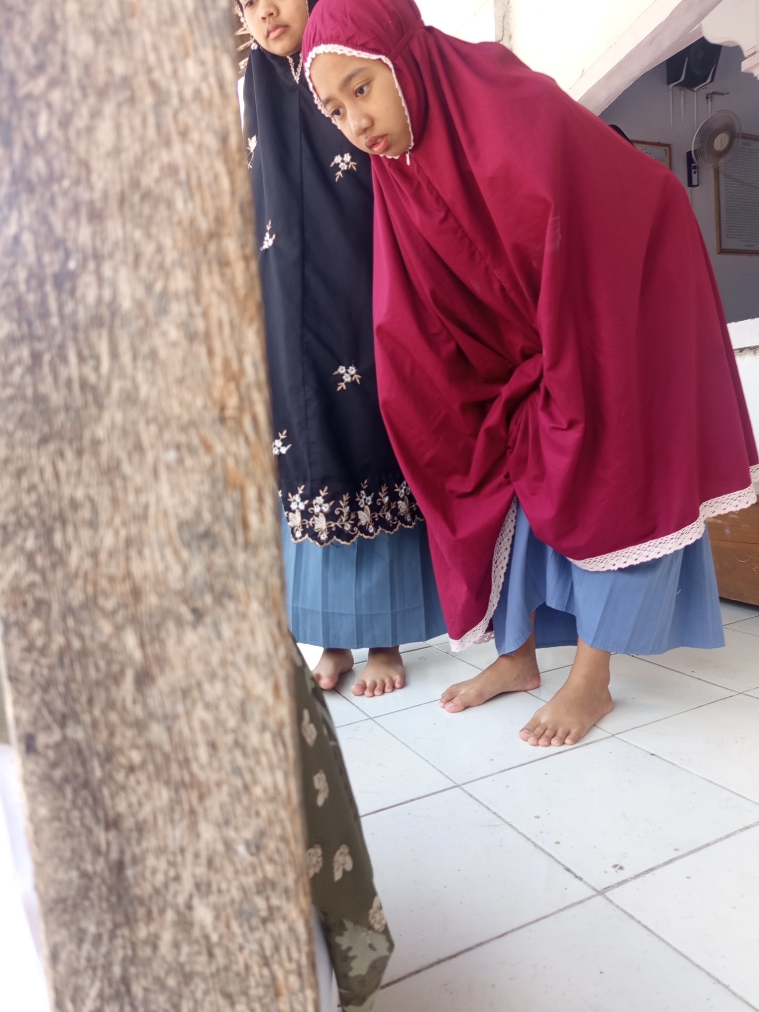 Two young Indonesian girls are standing on a white tiled floor. The girl on the right is wearing a maroon hijab and a blue skirt. She is looking down and to her left. The girl on the left is partially obscured by the girl on the right and a wooden post. She is wearing a black hijab and a blue skirt. Her face is not visible. The environment appears to be indoors, with a wall and ceiling visible in the background. There is a fan mounted on the wall and two framed pictures. The wooden post in the foreground is rough and weathered.