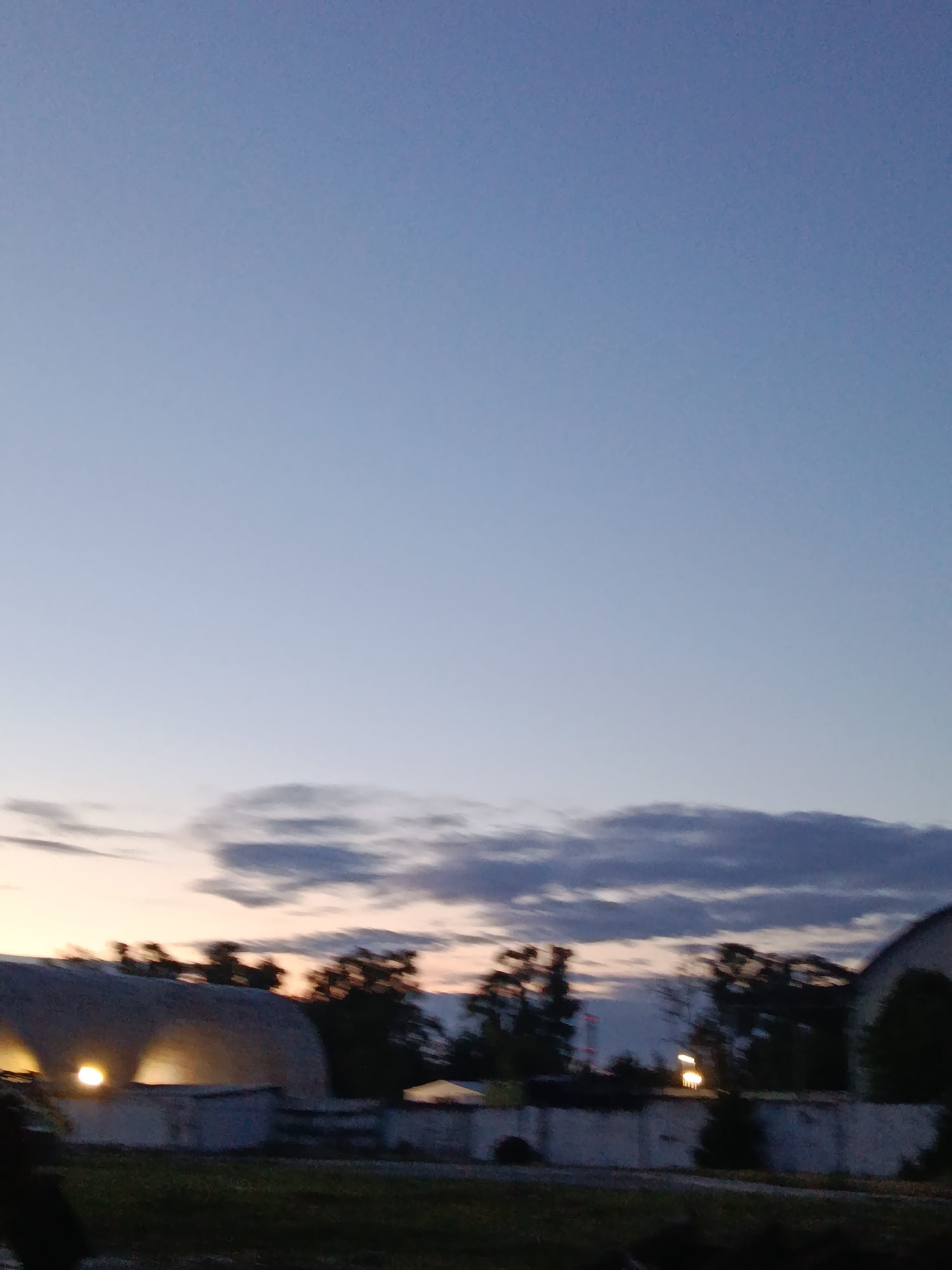 The image depicts a twilight scene over Svietlahorsk, Belarus. The sky is a gradient of pale blue and gray, with wispy clouds scattered across the horizon. In the foreground, several low-lying structures with rounded roofs are visible, partially obscured by trees. Artificial lights are illuminated within one of the structures, casting a warm glow. A concrete wall runs horizontally across the middle ground, separating the structures from a grassy area. The overall impression is one of a semi-rural or industrial area at dusk.
