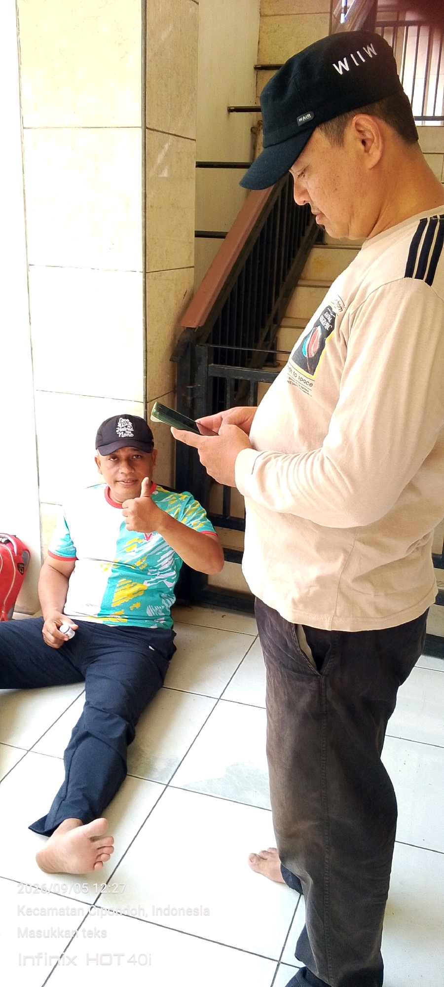 Two adult males are present. One male, wearing a black baseball cap and a light-colored long-sleeved shirt with a graphic on it, is standing and looking down at a mobile phone he is holding in his hands. The other male is sitting on the floor, wearing a black baseball cap, a multi-colored t-shirt, and dark pants. He is giving a thumbs-up gesture with his right hand. The scene appears to be outdoors, near a building with beige tiled walls and a staircase in the background. The floor is tiled with white squares. The timestamp on the image indicates the location is "Kecamatan Cipondoh, Indonesia."