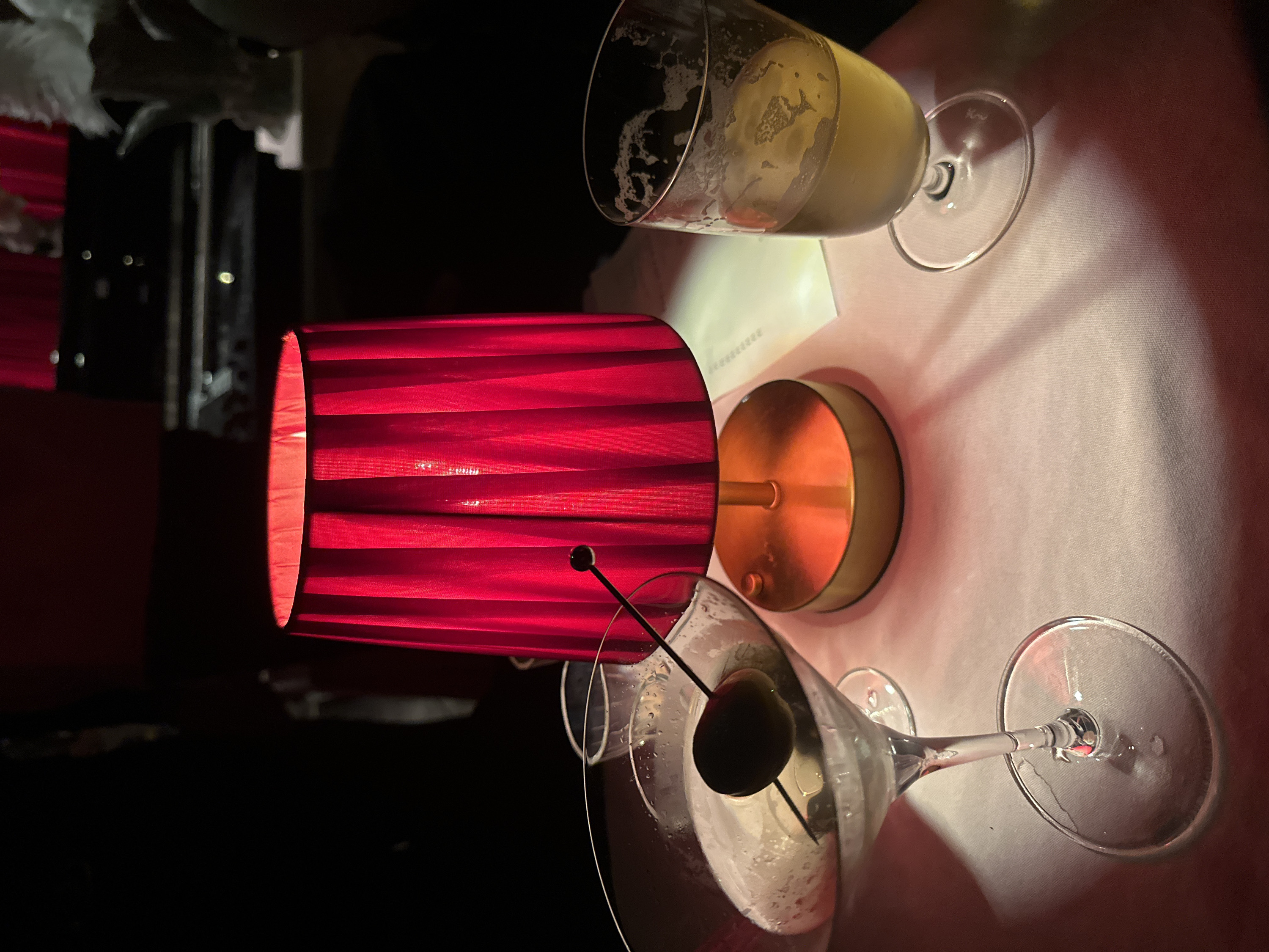A low-angle shot shows a red, pleated lampshade casting light on a white tablecloth. An empty champagne flute and a martini glass with an olive rest on the table. The martini glass is slightly closer to the viewer. The lampshade has a brass base. A dark, blurred background adds contrast to the scene. The lighting is dramatic, with strong shadows and highlights. A menu is barely visible under the flute.