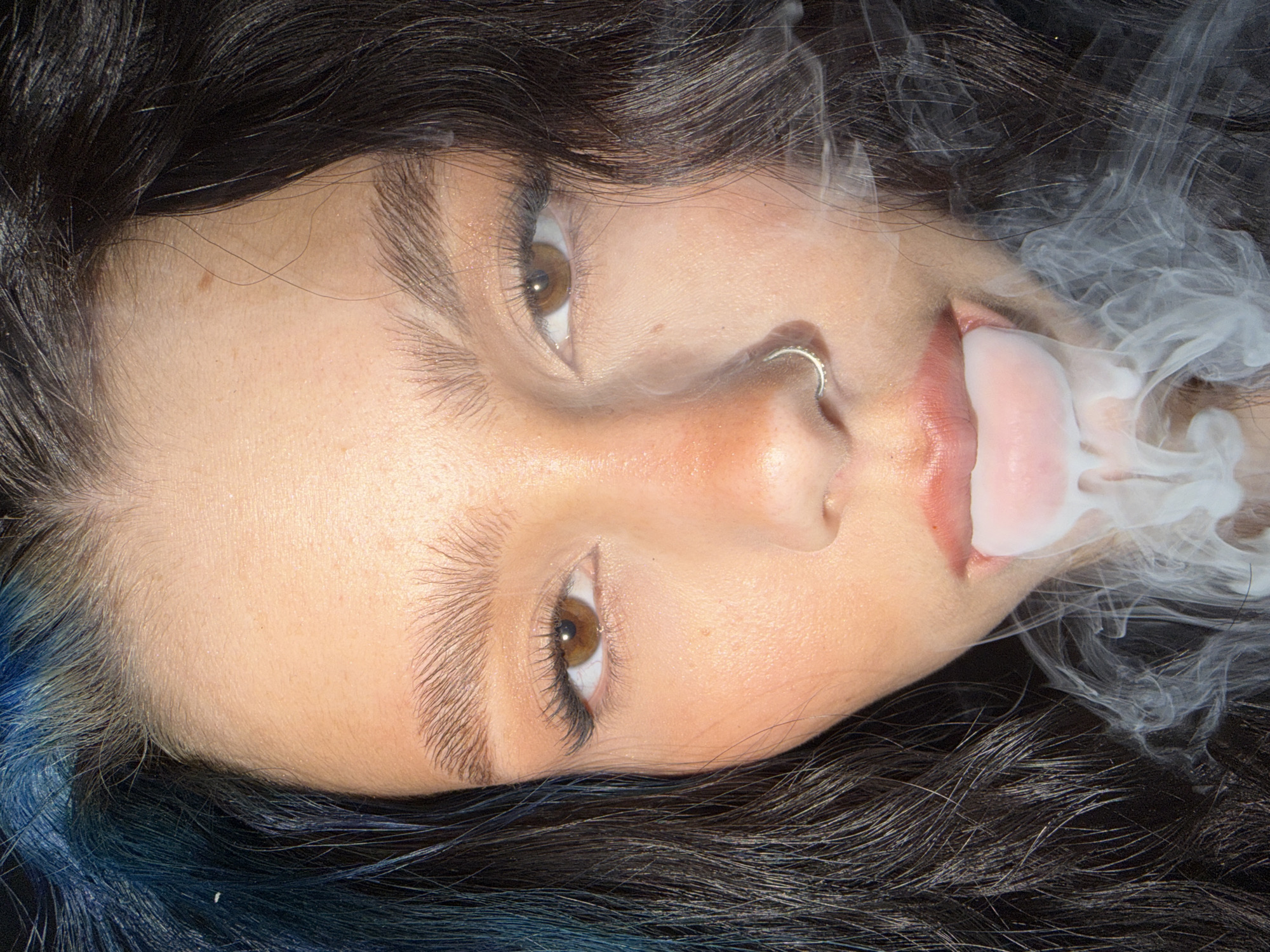 A medium shot depicts a woman lying down, with her head oriented towards the top left of the frame. She has fair skin, brown eyes, and dark brown hair with blue highlights near her left temple. A nose ring adorns her left nostril. The woman is exhaling white smoke, which obscures part of her mouth and disperses around her face and hair. The background is dark and indistinct. There is no information available in the image to indicate the location in the city of Khimki, Russia.