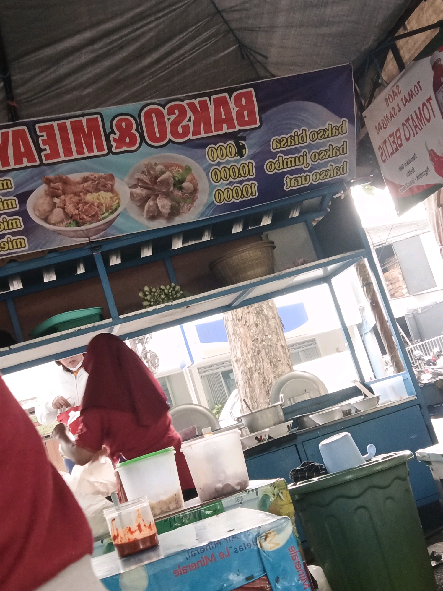 A street food stall in Kediri City, Indonesia, displays a banner advertising "BAKSO & MIE AYAM" (Meatball & Chicken Noodle Soup) with prices listed as 10.000.01 and 10.000.00, along with other illegible prices. Another sign to the right advertises "TOMATO BELTI" and "SARI" in red lettering. The stall has blue shelving filled with various food items and serving containers. A person wearing a red headscarf and apron is serving customers, and another person in a red shirt is partially visible in the foreground. In the background, a tree trunk and more stalls can be seen. The overall scene depicts a busy outdoor food vending operation.