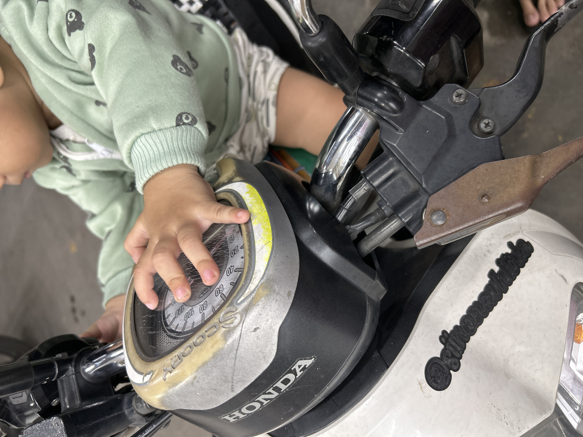 The image shows a close-up of a young child's hands interacting with the handlebars and dashboard of a Honda Scoopy scooter. The scooter's dashboard, displaying speed markings from 0 to 120 km/h, is partially obscured by the child's hands. The child is wearing a light green long-sleeved sweatshirt with a cartoon bear pattern and light-colored shorts. The scooter is white and black, with visible parts of the handlebars, brake levers, and a black sticker on the front panel that appears to read "Snow White". The scene suggests an informal setting, possibly a parking area or a home driveway in Depok, Indonesia, given the typical use of scooters in the region. The background is out of focus, showing a gray, textured surface consistent with pavement.