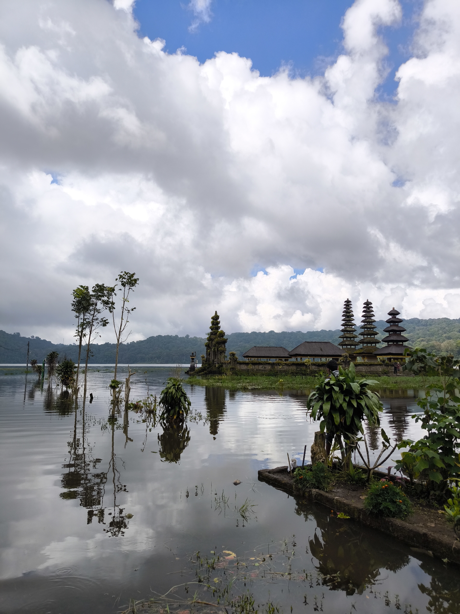 The image depicts a serene lake landscape in an unknown city in Indonesia. Dominating the mid-ground are structures with tiered, pagoda-like roofs, characteristic of Balinese temples. The structures are situated on the edge of the lake, reflecting in the calm water. In the foreground, there is vegetation growing from a small concrete platform. The lake is surrounded by lush, green hills in the background. The sky is filled with a mix of puffy white clouds and patches of blue, suggesting a partly cloudy day. The scene is still and peaceful, with the water acting as a mirror, reflecting the sky and the temple structures.