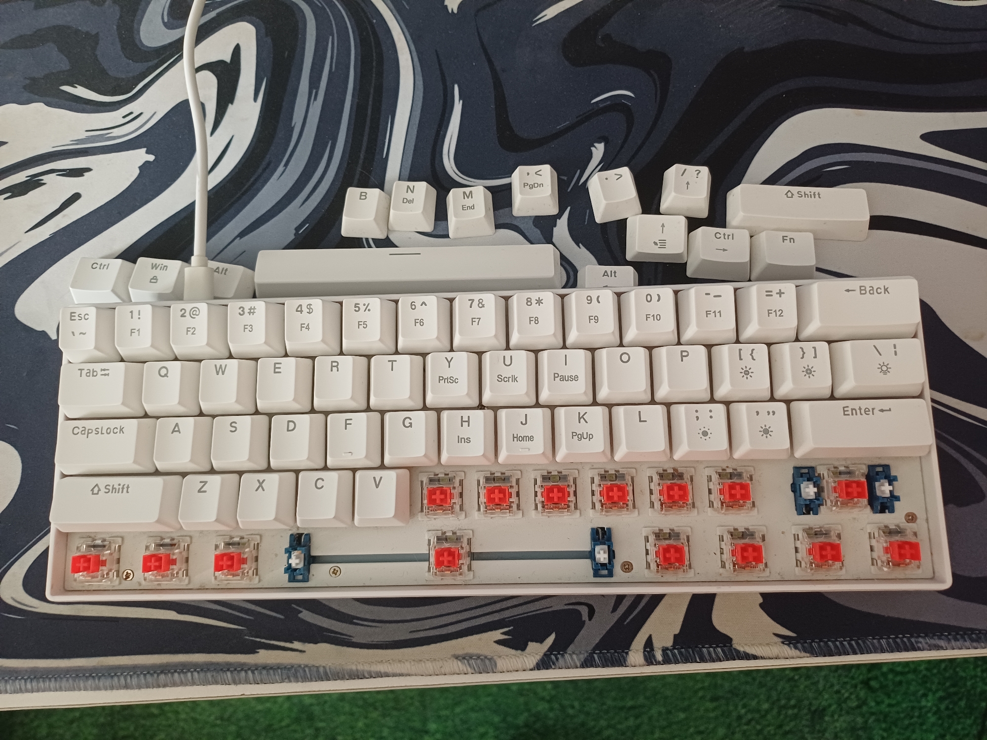 This media file shows a close-up, top-down view of a white mechanical keyboard situated on a desk with a marbled black and white desk mat. The keyboard is the primary subject, with its white keycaps and visible mechanical switches underneath some of the removed keycaps. Several keycaps are detached and scattered around the top section of the keyboard, including function keys, navigation keys, and letter keys. The visible switches appear to be red.
The scene is indoors, likely on a desk, as suggested by the keyboard and desk mat. There are no people visible in the image. The lighting appears to be neutral, suggesting it could be daytime or well-lit indoor conditions. The background is dominated by the abstract marbled pattern of the desk mat, which has swirling grey, black, and white tones.
Notable details include the text on the keycaps, such as "Ctrl", "Win", "Alt", "Esc", "Shift", and the function keys F1 through F12. Individual letter keys, numbers, and symbols are also visible. The presence of detached keycaps suggests a user might be customizing or cleaning the keyboard. The location context of Middlesbrough, United Kingdom, is not directly evident from visual cues within the image itself, but is provided as external information.