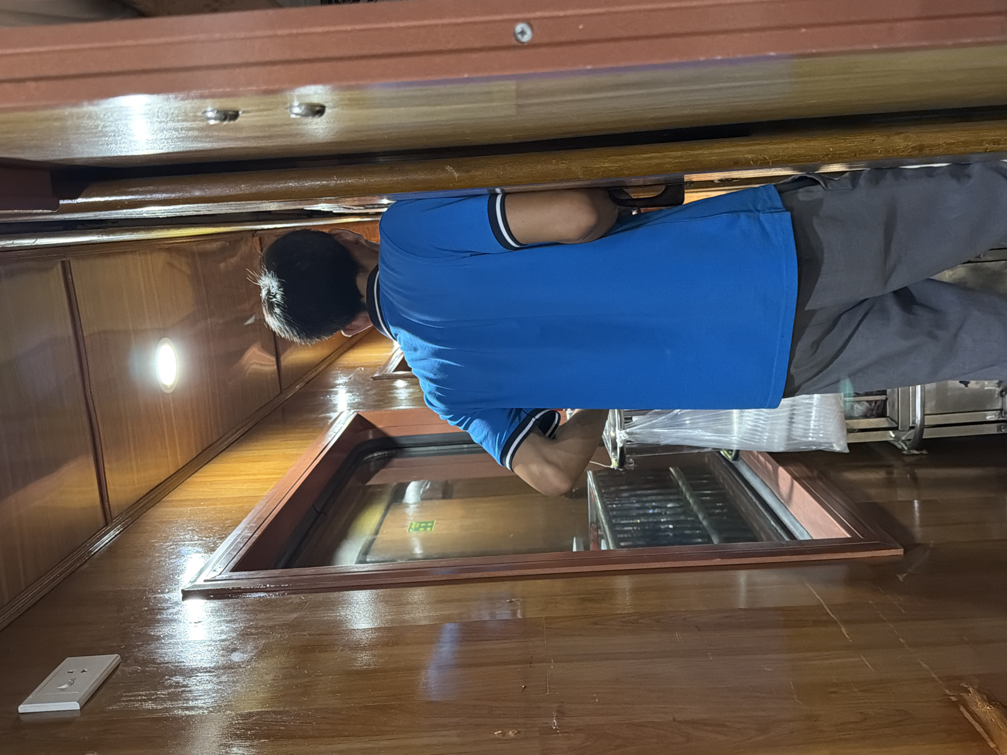 A person wearing a bright blue t-shirt and grey trousers stands in a narrow hallway lined with polished wooden walls. The hallway appears to be inside a train carriage, judging by the window and the narrowness of the space. A light fixture is visible on the ceiling, and a power outlet is mounted on the wall to the left. The person is facing away from the camera, looking towards a doorway or opening to the right. A plastic bag hangs from a rack to the left of the person. The environment suggests an interior setting with a warm, polished wood aesthetic. No specific location in Da Nang, Vietnam can be determined from the image.