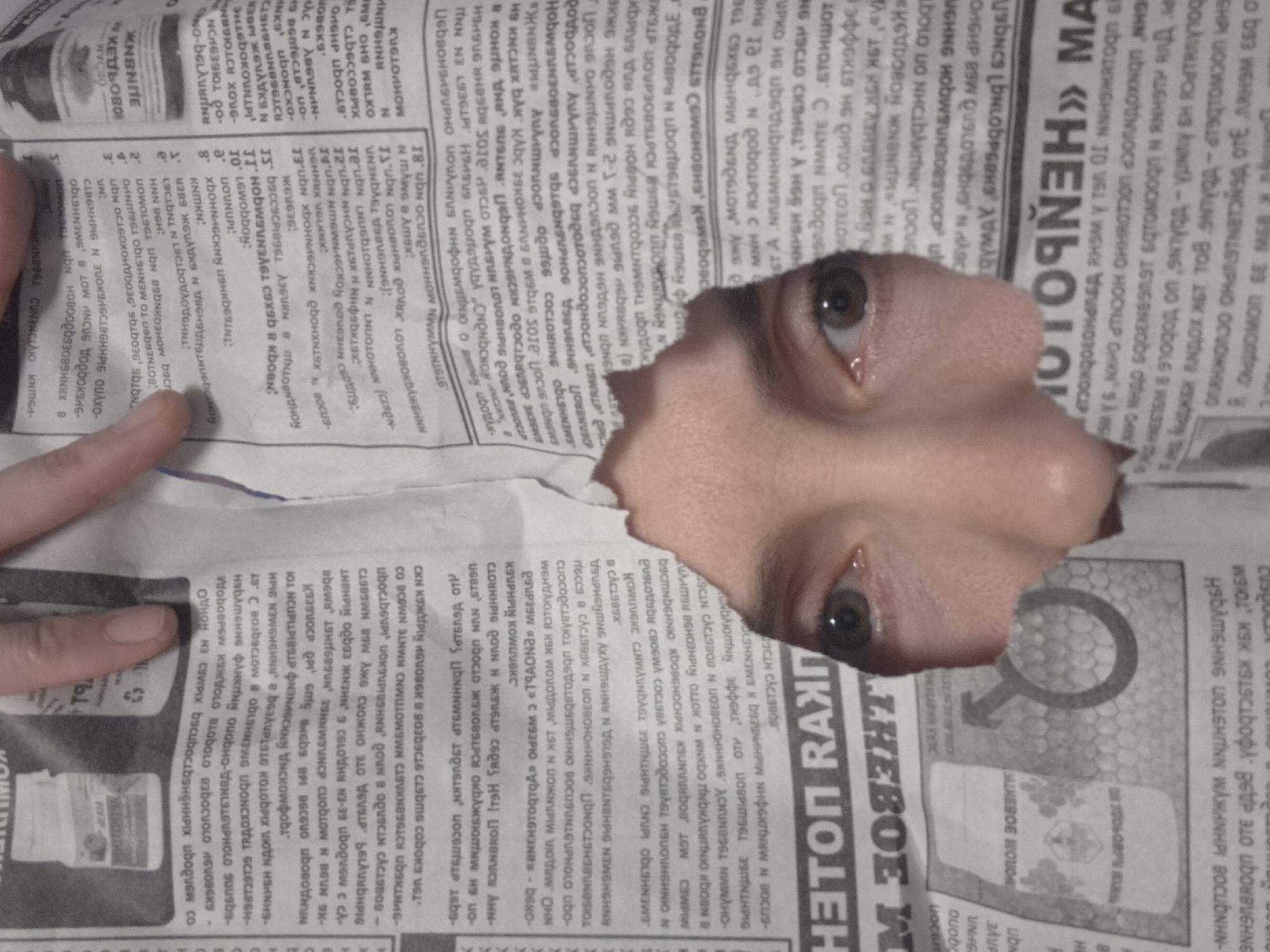 A person's face peers through a hole torn in a newspaper. The tear is jagged, revealing the skin around the eyes, nose, and part of the cheek. The newspaper is in black and white, filled with dense text in a Cyrillic alphabet. A hand with light skin and manicured fingernails holds the newspaper. The text is unidentifiable and the language is unknown. The location is presumably indoors. The context and relationship between the face and the newspaper are unclear.