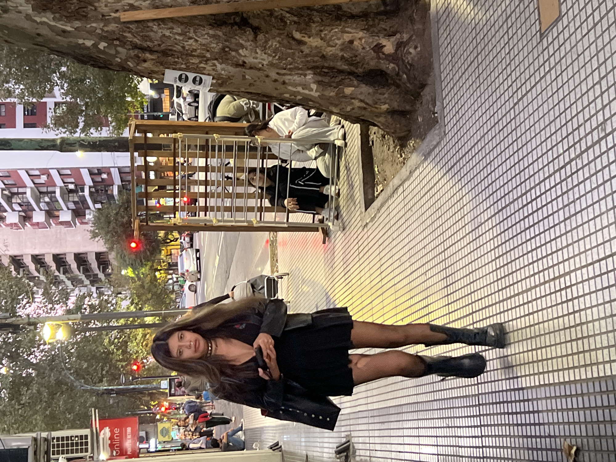 This media file depicts an urban street scene in Buenos Aires, Argentina, likely during the evening or dusk due to the illuminated streetlights and ambient light.
In the foreground, a young woman with long dark hair stands on a tiled sidewalk, looking directly at the camera. She is dressed in a black ensemble, including a dress, a leather jacket, fishnet stockings, and dark boots. A choker adorns her neck, and she holds a dark-colored phone in her hands. A black bag or jacket is draped over her arm.
Behind the woman, beneath a large tree trunk, a wooden structure serves as a seating area or shelter. Two individuals are visible within this structure: one wearing a white top and light trousers, and the other in dark clothing. A white sign with two black human figures (one seemingly male, one female) and a partially visible white drone are also present in this area.
The background features multi-story buildings with numerous windows, indicative of a residential or commercial district. Several streetlights are on, and red traffic lights can be seen along the street, suggesting vehicular traffic. Other pedestrians are visible further down the street. To the lower left, a red sign with white text reads "online" and "Punto de Retiro" (pickup point). The sidewalk features a repeating pattern of small, light-colored square tiles. The overall scene captures a moment of typical city life as evening settles.