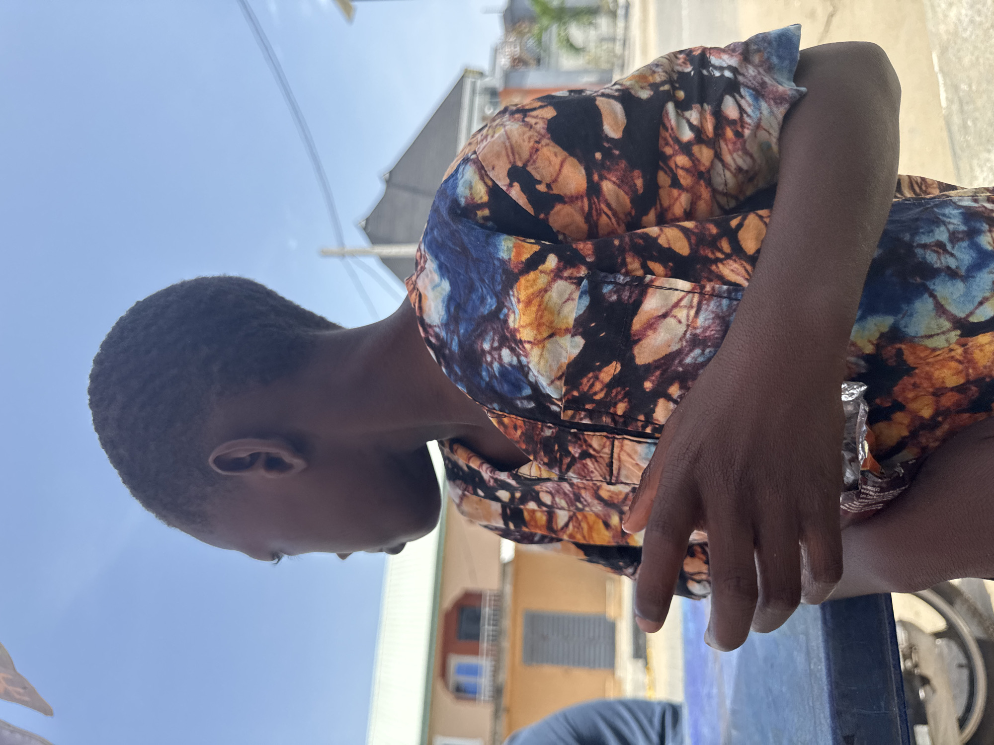A young dark-skinned person with short hair is sitting at a blue table outdoors, likely in Badagry, Nigeria, based on the architecture and clothing style. The person is wearing a patterned shirt with a mix of black, orange, and blue colors. The person's head is turned down, so only their profile is visible. A partial view of a building with a green roof and a small window with blue trim is in the background. A shiny metallic object and a tire are on or near the table. Overhead, the sky is visible.
