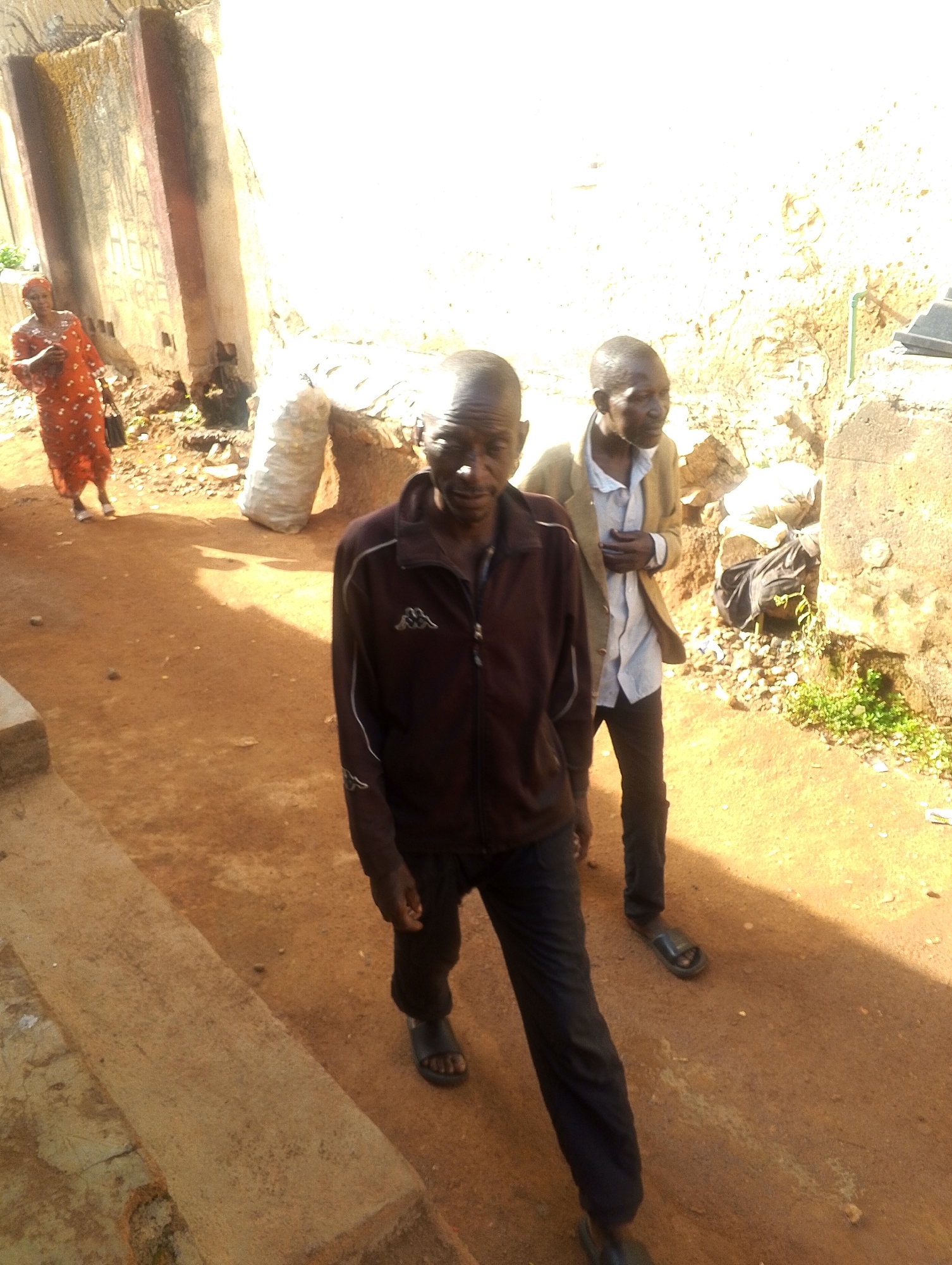 The image shows an outdoor scene in Jos, Nigeria. There are three individuals present. The main subject is a slender, older man wearing a dark brown zip-up jacket, dark pants, and sandals, who is walking towards the camera. Slightly behind him to the right, another older man is wearing a light blue shirt, a tan jacket, dark pants, and sandals. A woman in an orange patterned dress is walking away from the camera in the background to the left. The ground is an orange-red color. The backdrop is made up of structures with weathered and stained walls, and a large sack is visible in the background. Vegetation is observed on the right side of the frame. The lighting suggests it is daytime, and shadows are visible.