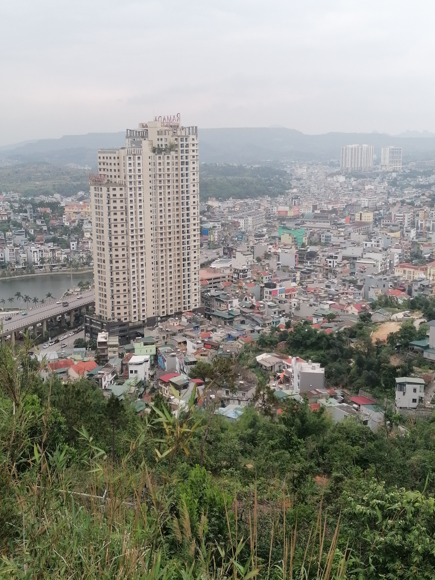 The image depicts an elevated view of Hong Gai Ward in Vietnam, characterized by dense urban development and a hazy atmospheric perspective. A prominent, multi-story hotel, labeled "RAMADA," stands out as a central architectural feature. A bridge traverses a body of water, connecting different sections of the urban landscape. The foreground contains a mix of dense vegetation, suggesting a position on a hillside or elevated ground. The overall tone is muted, potentially indicating early morning or overcast conditions. The dense collection of buildings and infrastructure suggests a populated area with diverse architectural styles. In the distance, rolling hills are visible, adding to the depth of the scene.