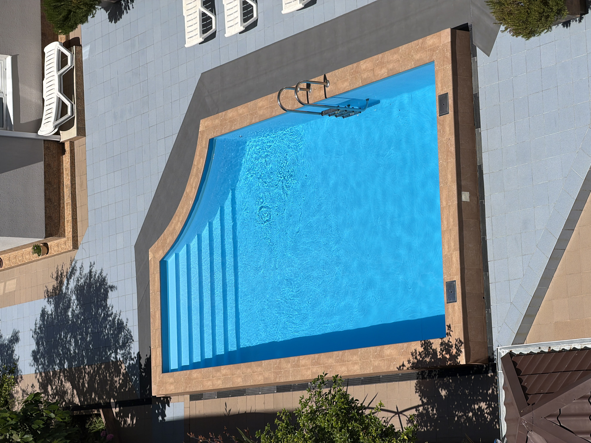 The image shows an aerial view of an outdoor swimming pool area. The pool is rectangular with rounded corners and has a built-in set of stairs. The water is a vibrant blue. The pool is surrounded by a tiled patio. To the right of the pool, there are several white lounge chairs. In the background, there is a building and some landscaping. The image appears to have been taken on a sunny day, as indicated by the shadows cast by trees. The location is identified as Sirius, Russia.