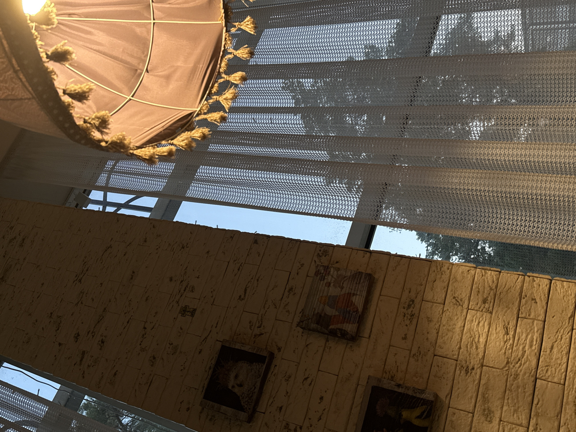 The image depicts an interior scene with a brick wall on the left and a window with sheer curtains on the right. A lampshade with tassels hangs in the upper right corner. Several small framed pictures are affixed to the brick wall. The lighting suggests it is either early morning or late afternoon. The scene is set indoors, likely a residential or commercial space. There is no indication of the city of Abinsk, Russia within the image itself.