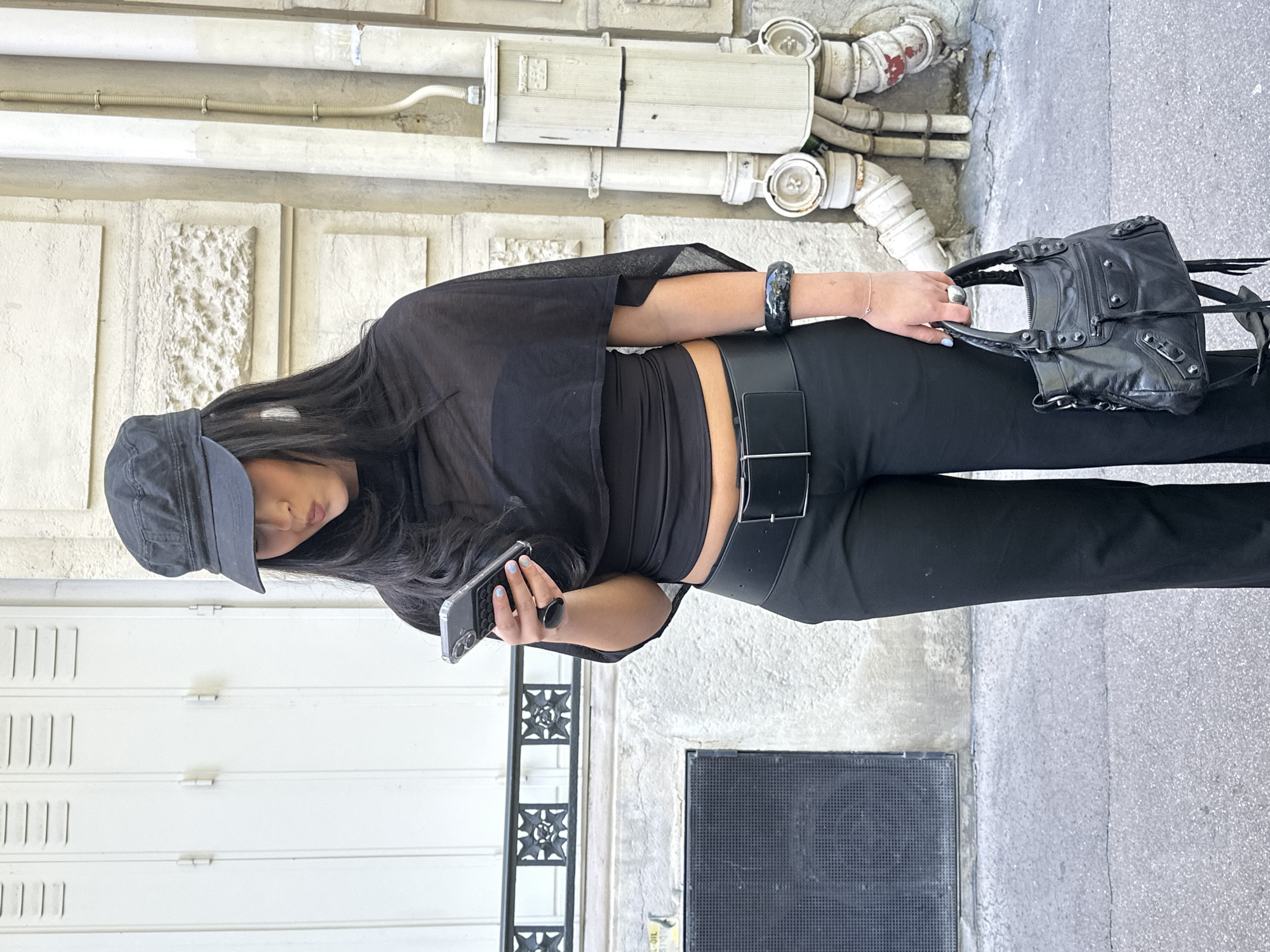 A medium eye-level shot depicts a person holding a black bag. The person is wearing a black brimmed cap, a black shirt, and black pants with a wide black belt. They are holding a phone with a black case in their left hand. A black bracelet is on their right wrist. The background features a wall, an electrical box with various white pipes, a gated area, and a ground-level gray grating. Based on the architecture, the image appears to be taken in Paris, France.