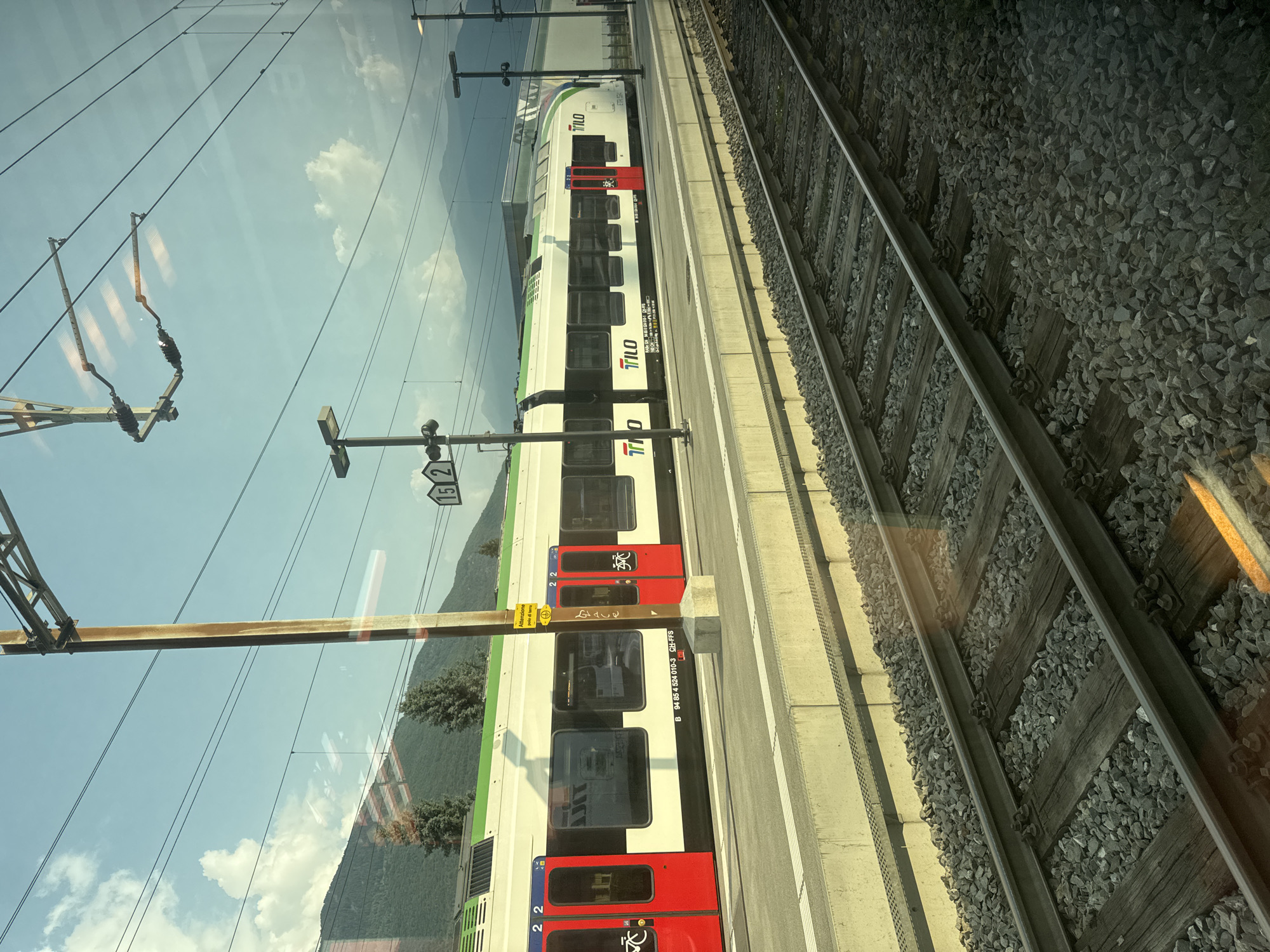 The image captures a train at a station in Bellinzona, Switzerland, on a bright, partly cloudy day. The train, with white and green livery, displays the "TILO" logo and appears to be a regional train service. Red doors with bicycle symbols are visible along the side of the train. The train is stopped at a platform constructed with concrete and edged with yellow lines. Train tracks, laid on ballast, run in the foreground, indicating this is a view from another train or a pedestrian area beside the tracks. Overhead electrical lines and poles for the railway infrastructure are prominent in the upper portion of the image. In the background, lush green mountains rise against a blue sky dotted with white clouds. A yellow warning sign with Italian text, "Attenzione polo di terra," is affixed to a pole near the train. The overall scene suggests a typical day at a Swiss railway station, surrounded by the natural beauty of the Ticino region.