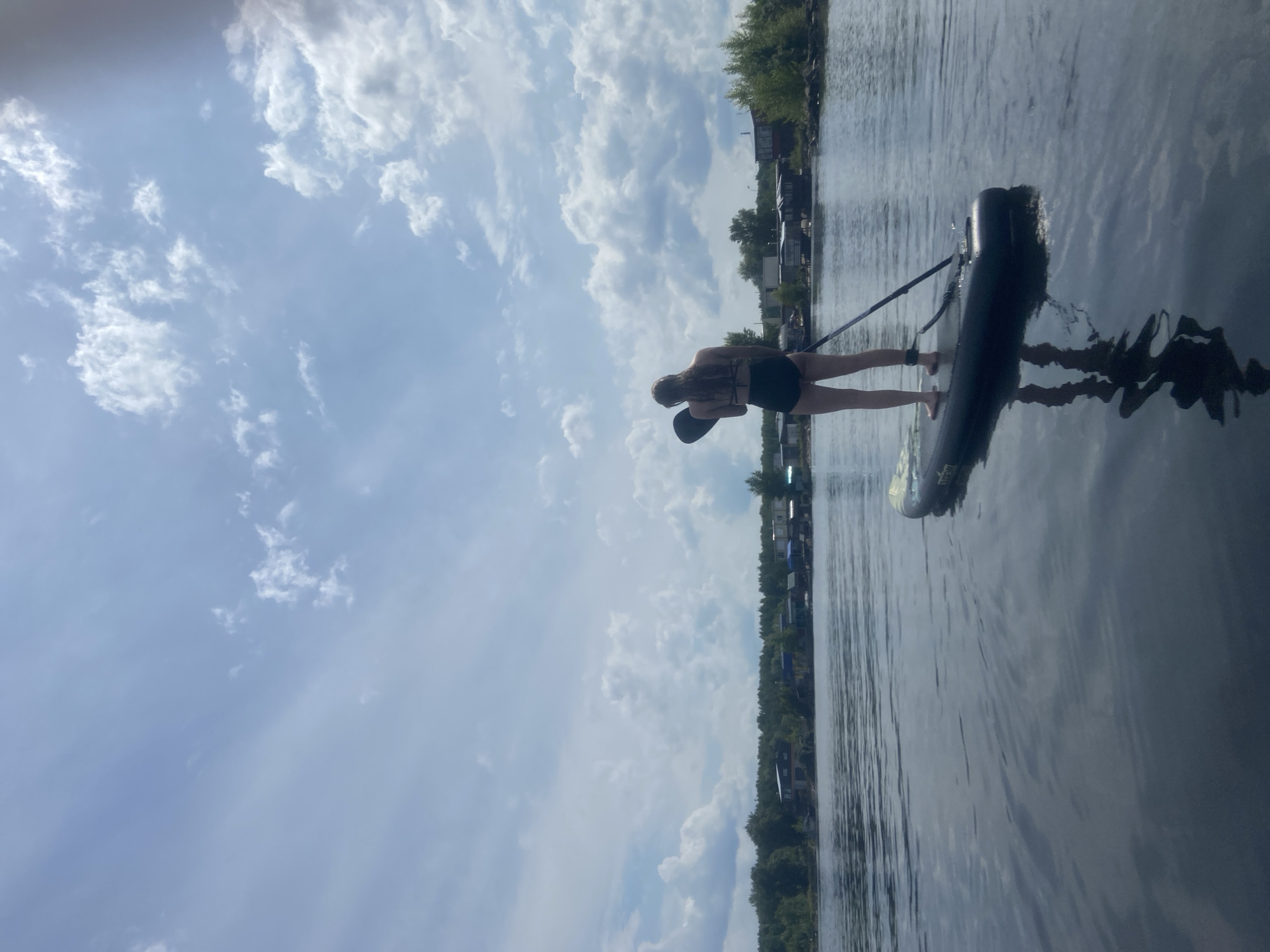 A woman stands on a paddleboard in the water, facing away from the camera. She is wearing a black bikini and has long, light-colored hair. She is holding a paddle. The paddleboard is black and appears to be inflatable. The water is calm and reflective, showing the sky and the woman's reflection. In the background, there is a shoreline with trees and several buildings, which appear to be small houses or cabins. The sky is partly cloudy with scattered white clouds against a blue background. The overall impression is a serene outdoor scene, likely a lake or river in a rural or semi-rural area. This scene is set in the city of Chaikovsky, Russia.