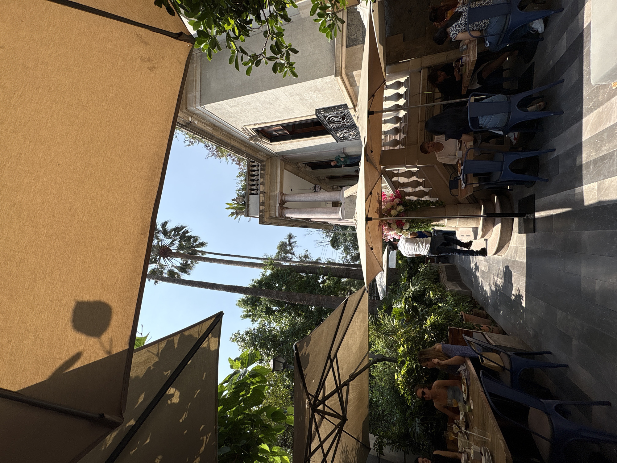 The image shows an outdoor dining area in Mexico City, Mexico. Several people are seated at tables, enjoying food and drinks under large beige umbrellas. The dining area is adjacent to a building with ornate architectural details, including columns, a balcony, and decorative ironwork. Tall palm trees are visible in the background, adding to the tropical feel of the scene. Sunlight casts shadows across the dining area. Some people are seated, others are standing. Some people are looking at their phones, others are conversing with their companions.