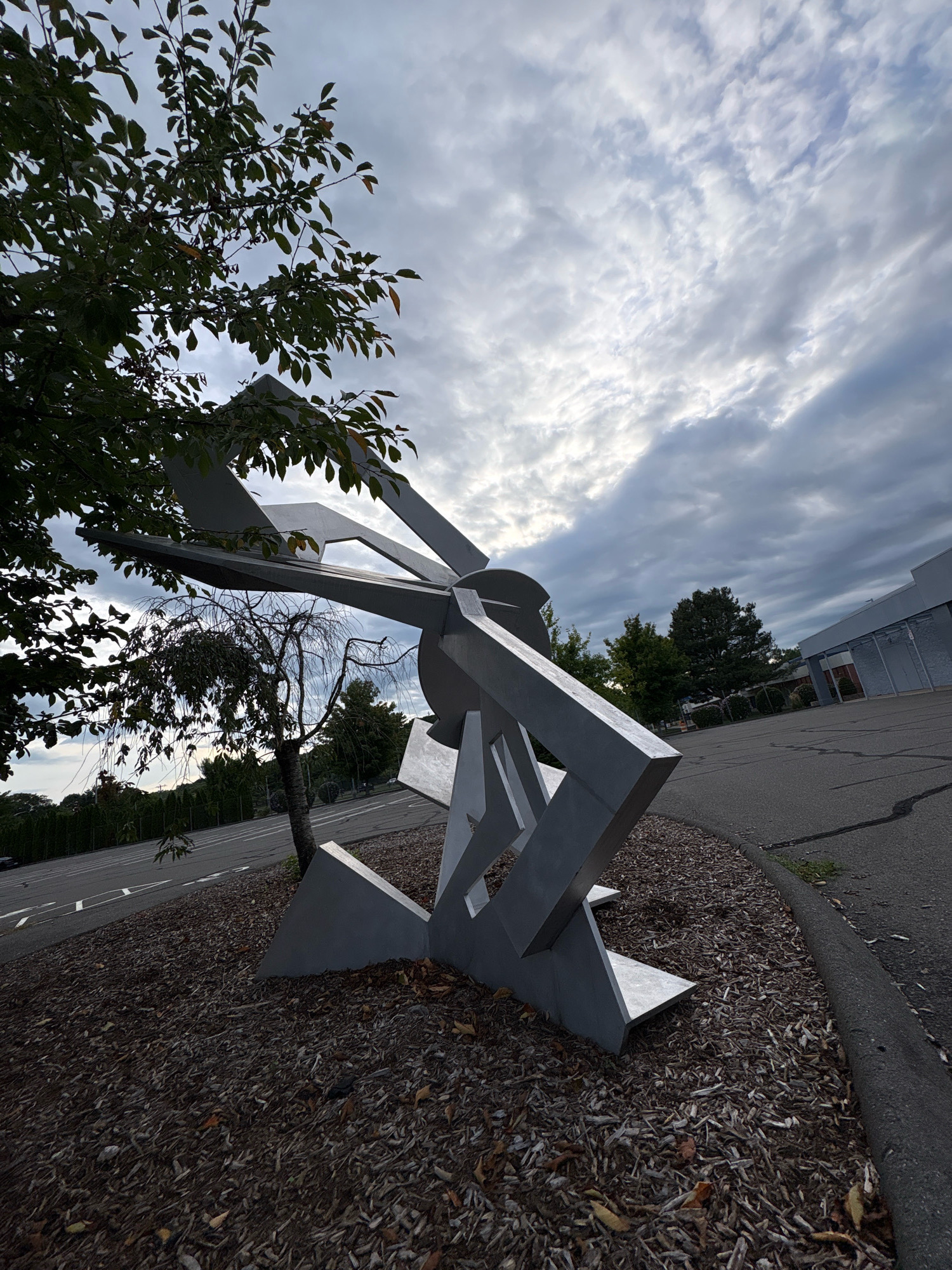 The image depicts an abstract metal sculpture in an outdoor setting, possibly a park or public space, with a cloudy sky overhead. The sculpture is geometric and angular, constructed from light-colored metal and appears to be a significant size, dominating the foreground. To the left of the sculpture, a tree with green leaves partially obstructs the view, while another smaller, bare tree is visible behind it. The ground around the sculpture is covered in mulch and fallen leaves. In the background, a paved area with road markings is visible, suggesting it might be a parking lot or street. Further in the distance, there are trees and a building with a light-colored facade. The overall atmosphere is overcast and the lighting is muted. The specific location in the city of Windsor, United States of America, is not identifiable from the visual information provided. There are no people or animals present in the image.