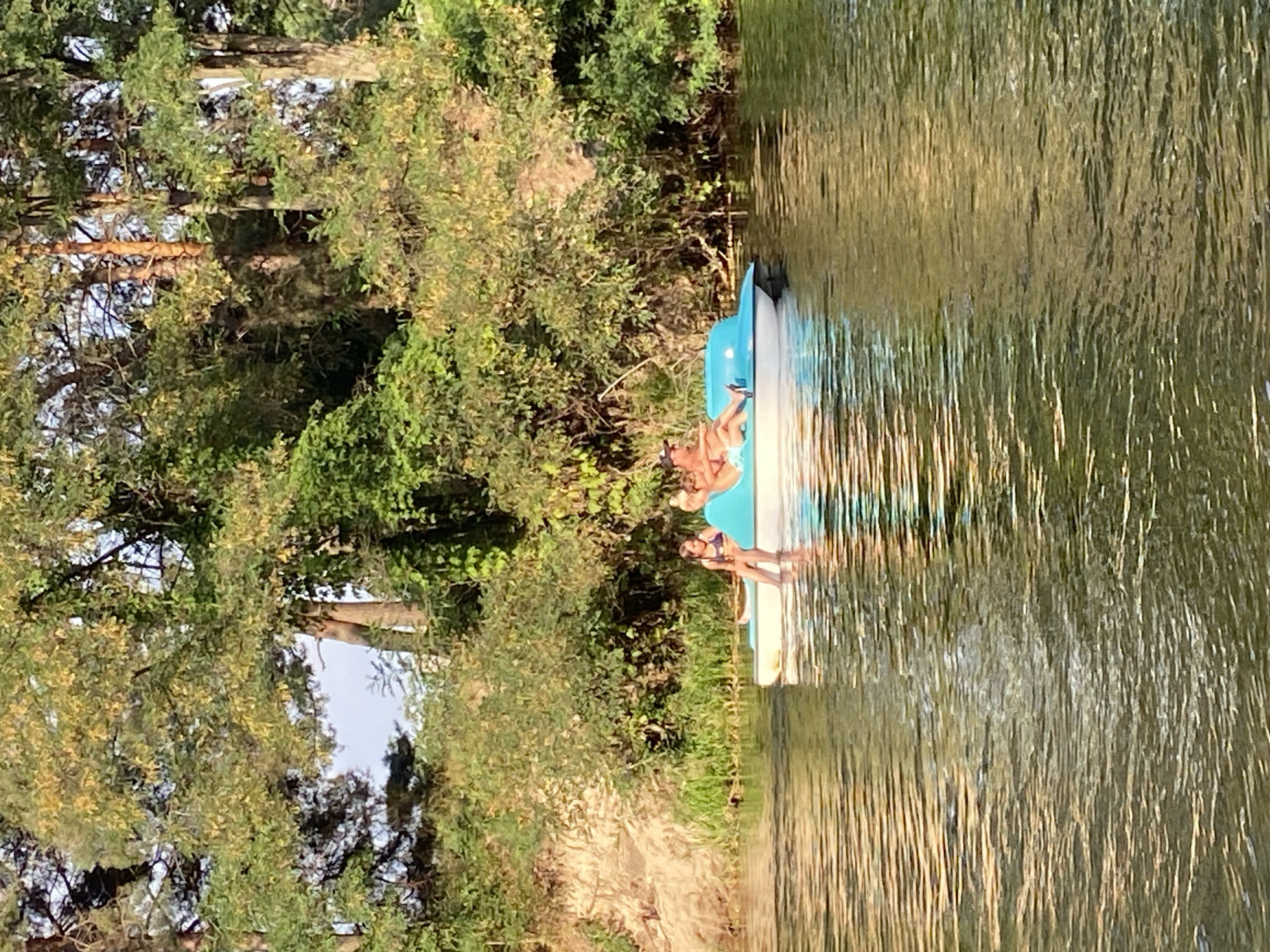 A wide shot captures a pedal boat on a calm body of water, likely a river or lake, surrounded by lush green trees and vegetation that line the bank. The sun is casting warm light, creating reflections on the water's surface. On the pedal boat, two individuals are seated. A person with light-colored hair, possibly a child, is seated towards the front of the boat, wearing a dark-colored swimsuit. To their right, an adult is seated and appears to be wearing a light-colored top and dark bottoms. The boat is blue and white and has a pedal mechanism visible. The bank of the water features a sandy embankment with dense green foliage above it. The overall scene suggests a peaceful outdoor recreation activity on a sunny day in Semianivka, Ukraine.
