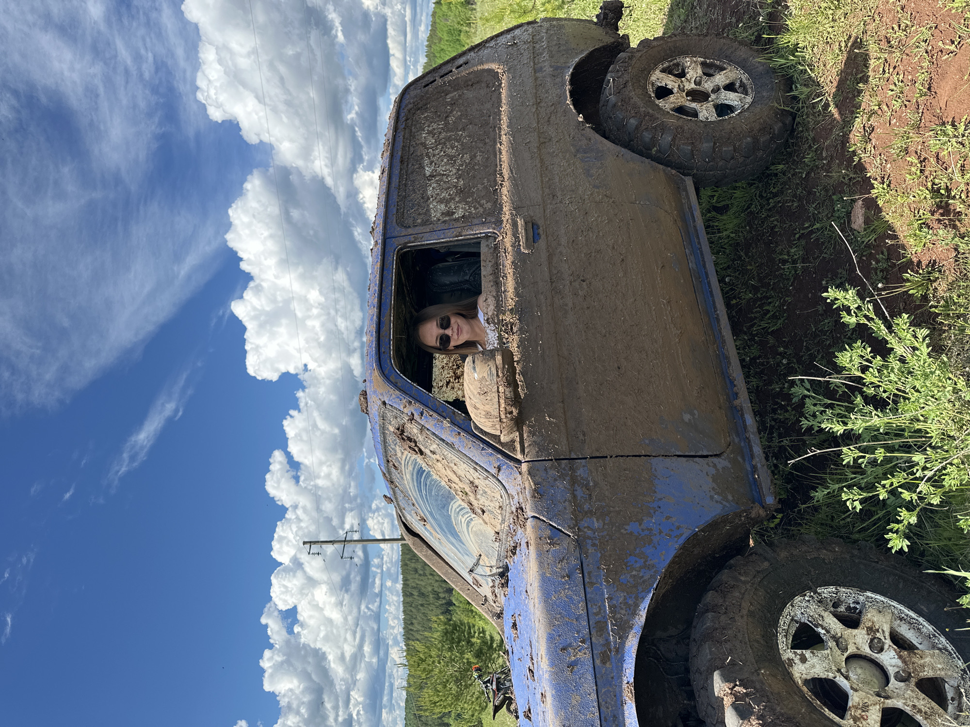 A blue, mud-covered SUV is positioned at an angle on a dirt and grass incline. The driver, a female wearing sunglasses and a white shirt, is visible through the driver-side window. The sky features a blue expanse with scattered white clouds and power lines. The scene appears to be located in a rural area near Ilyinsky, Russia, due to geographic inference.