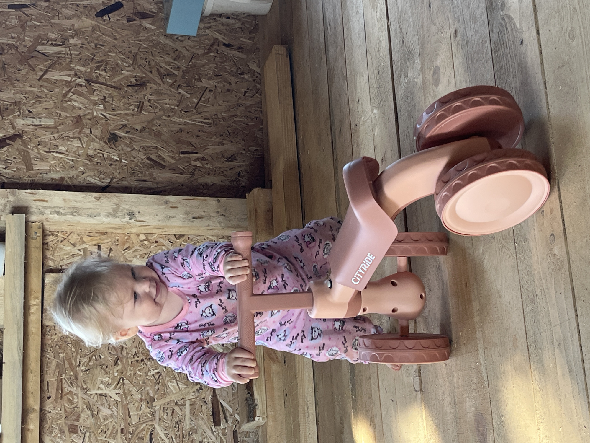 An infant with blonde hair, wearing pink pajamas with a pattern of cartoon animals, stands behind a pink children's ride-on toy. The infant's hands are on the handlebars of the toy, and they are looking down and to their right with a slight smile. The ride-on toy has "CITYRIDE" printed on its side. The background consists of wooden plank flooring and a wall made of particle board. The scene appears to be indoors, possibly in a workshop or unfinished room. The city of Fokino, Russia, is not depicted in this image.