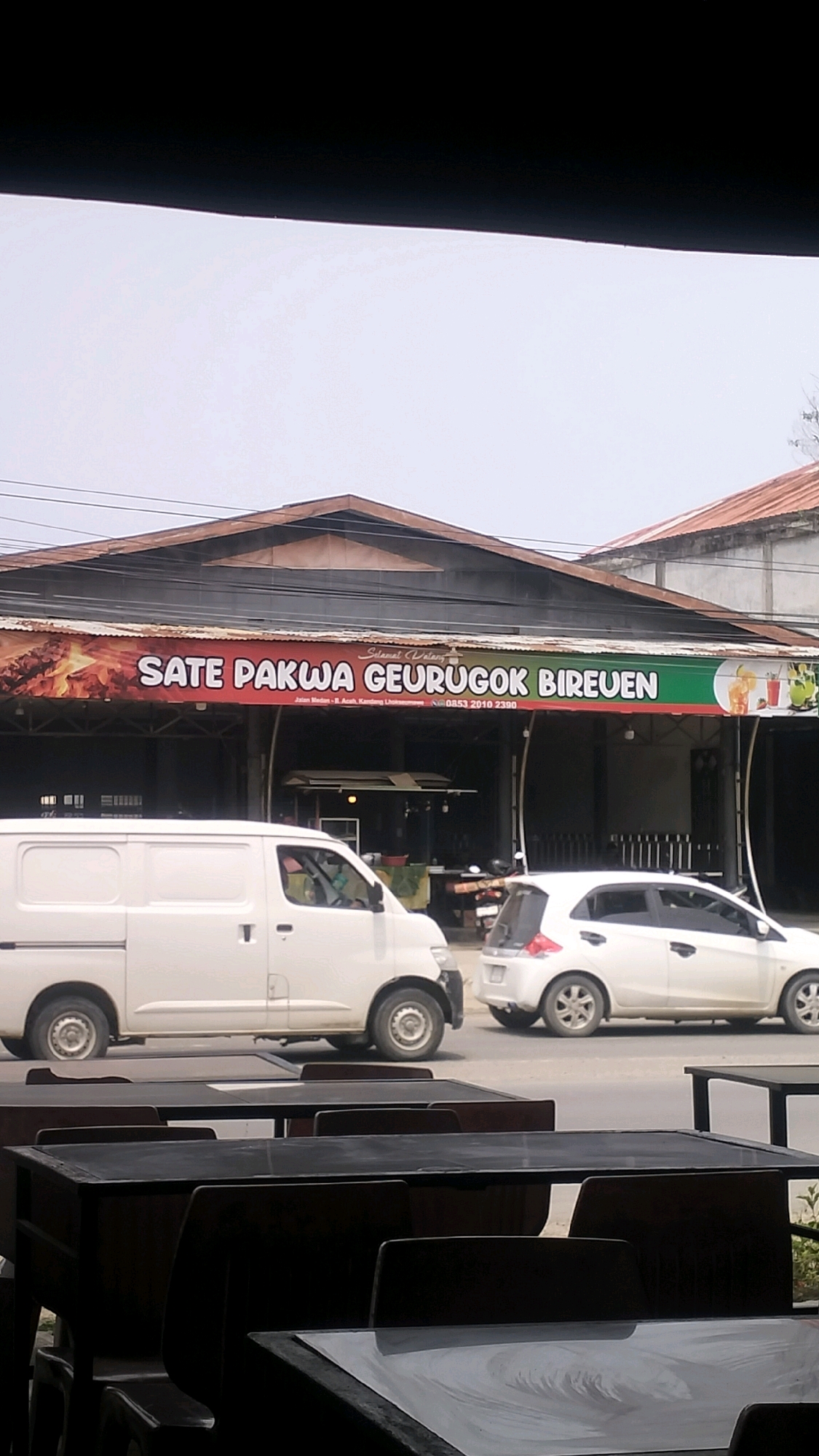 The image shows an outdoor dining area with tables and chairs in the foreground, facing a street. Beyond the tables, a building with a prominent banner is visible. The banner reads "Sate Pakwa Geurugok Bireuen" and includes the text "Selamat Datang" (Welcome), a street address "Jalan Medan - R. Aceh, Kandeung, Uleekareng", and a phone number "0853 2010 2390". Two white vehicles, a van and a car, are parked on the street in front of the building. The building appears to be a restaurant or food establishment. The sky is overcast. The scene depicts a typical urban streetscape in Lhokseumawe, Indonesia, with commercial activity and traffic.