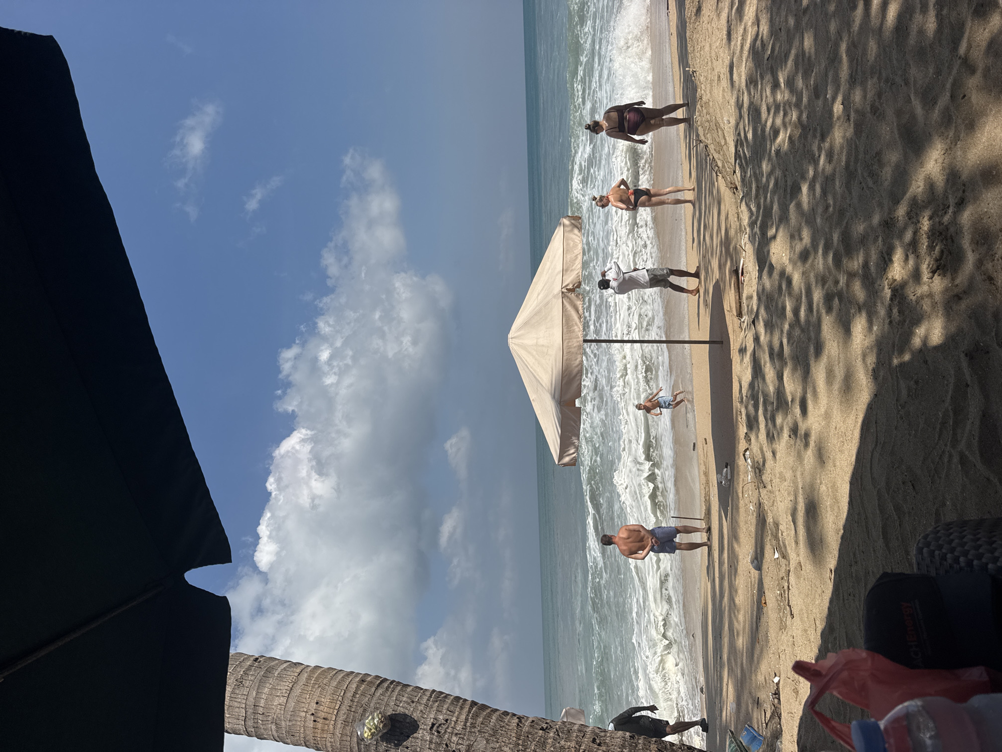 In Kuta, Indonesia, a crowded beach scene unfolds under a bright, partly cloudy sky. The foreground is dominated by sand, with the dark shadow of an overhead umbrella cast across it. To the left, the rough, textured trunk of a palm tree stands tall. In the middle ground, a line of people stands on the wet sand near the ocean's edge. A man in blue shorts faces away from the camera, gazing at the crashing waves. Nearby, a child in swimming trunks runs towards the water. To the right, under a beige umbrella, a man in shorts and a white shirt holds up a camera, seemingly taking a photo of two women in bikinis. The woman on the left wears a black bikini, while the woman on the right wears a dark red bikini. The ocean is active, with white-capped waves rolling towards the shore. Scattered debris, including plastic bags and bottles, litters the sand near the water.