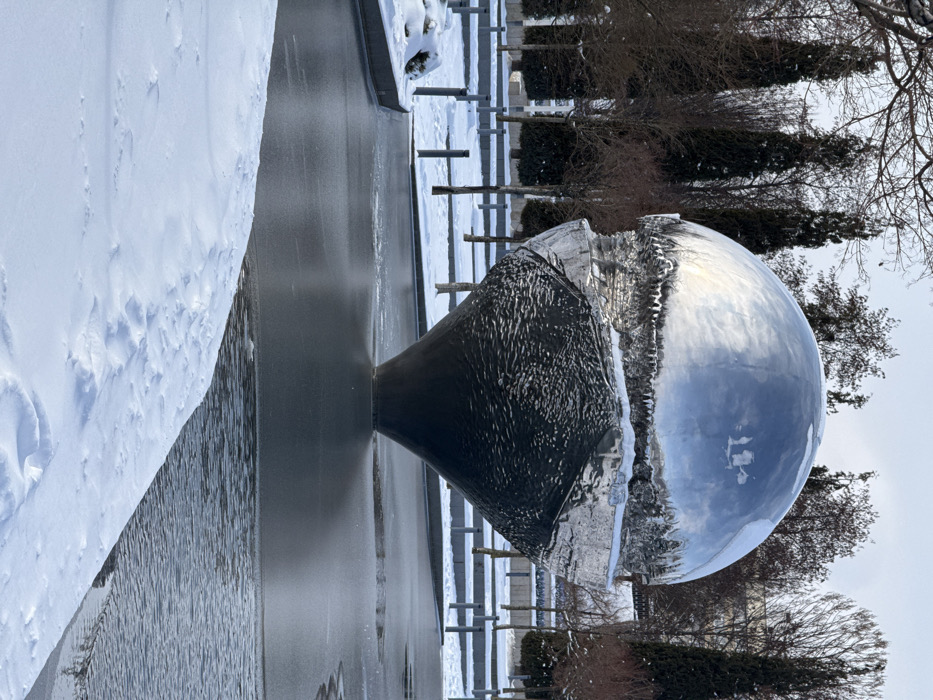 The image depicts a winter scene featuring a large, spherical art installation situated within a partially frozen body of water in a landscaped urban park in the city of Plodorodny, Russia.
The central object is a prominent sculpture, roughly 5-6 meters tall, composed of two distinct halves. The upper hemisphere is highly polished, reflective metal, possibly chrome, which mirrors the pale, overcast sky and the silhouettes of surrounding trees. A noticeable layer of white snow and numerous icicles are present along the sculpture's equator, marking the transition point between the two halves. The lower half of the sculpture is a dark, textured, conical form, appearing to emerge from or be supported by the water.
In the foreground and surrounding the sculpture is a body of water, partially covered with dark, reflective ice patches, while some areas of open water show slight ripples. Snow blankets the banks adjacent to the water and extends across the ground surfaces in the background. The background features a mix of bare deciduous trees and tall, dark evergreen trees, likely cypress or thuja, indicating a cold climate. Paved pathways or pedestrian areas, also covered in snow, are visible further back, interspersed with dark, short bollard-like structures. No individuals are present in the scene, and no specific actions or interactions are depicted.