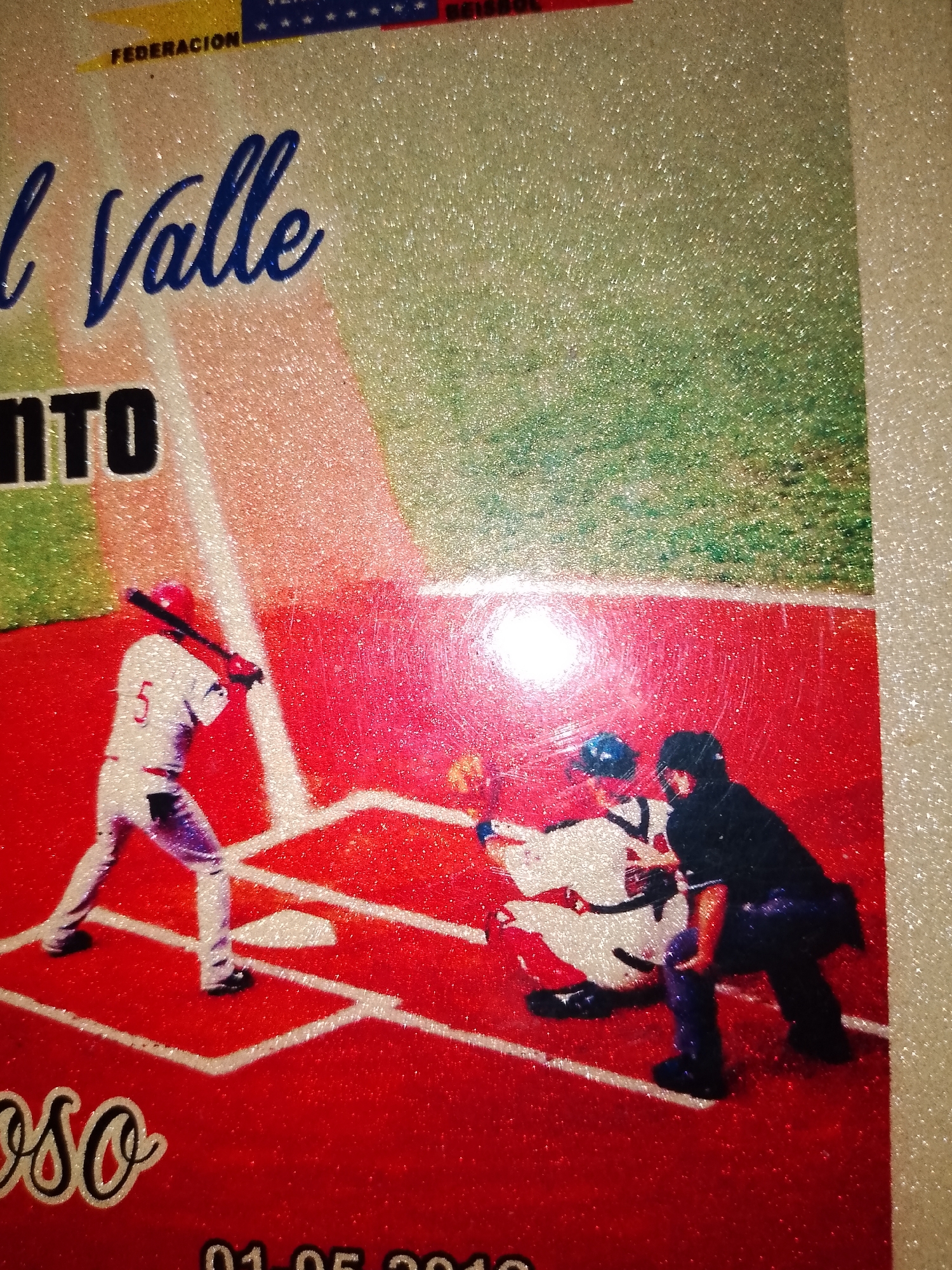The image shows a close-up of a poster depicting a baseball game. In the foreground, a batter in a white uniform with the number "5" on his back is swinging at a pitch. A catcher in a blue helmet and a dark uniform is crouched behind home plate, and an umpire in a black uniform is also visible. The background shows a baseball field with green grass and red dirt. Text in Spanish is visible at the top of the poster, including "FEDERACION" and "l Valle". There is also text at the bottom that appears to be a date: "01-05-2010". The poster is glittery, suggesting it may be a decorative item or part of an event. The location mentioned, "Santa Ana, Venezuela," is not directly depicted in the image itself but is a contextual piece of information about the poster's origin or purpose.
