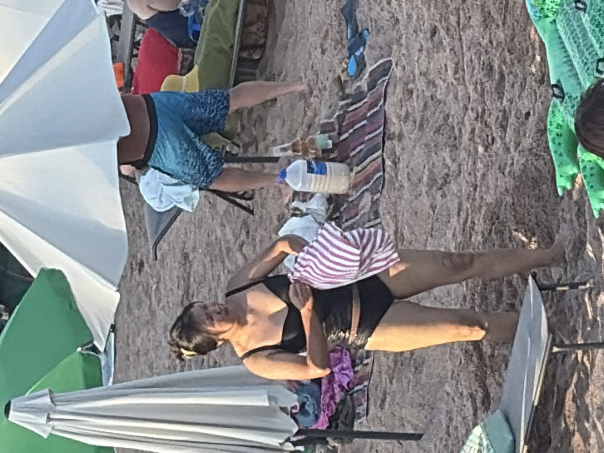 The image shows a sandy beach with beach umbrellas, lounge chairs, and people. In the foreground, a woman wearing a black bikini is holding a striped towel. She is looking to the right. Behind her, a person wearing blue patterned swim trunks is standing under a white umbrella. Several beach chairs and colorful towels are visible in the background. A large plastic water bottle and a glass bottle are on the sand. The scene is outdoors on a beach.