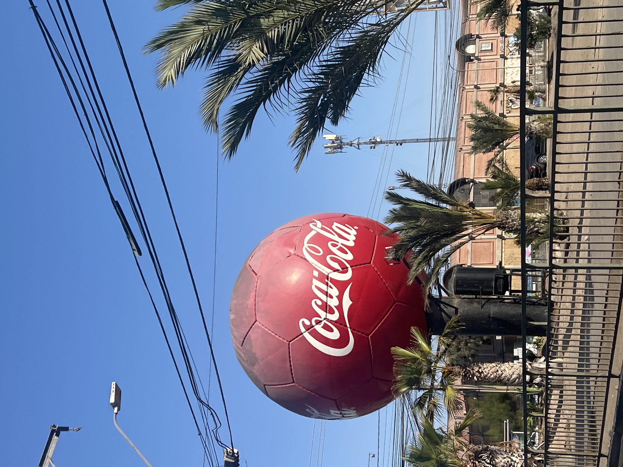 A low-angle, medium shot shows a large, spherical Coca-Cola sign in Arica, Chile. The sign is red and white and appears to be made in the design of a soccer ball. The sign is located next to a black metal structure and several palm trees. A brick building is behind the palm trees and a black sedan is parked in front of it. Power lines are visible in the upper left portion of the image and a communication tower is located further away in the background. The sky is clear and blue. A metal fence is visible on the right edge of the frame.