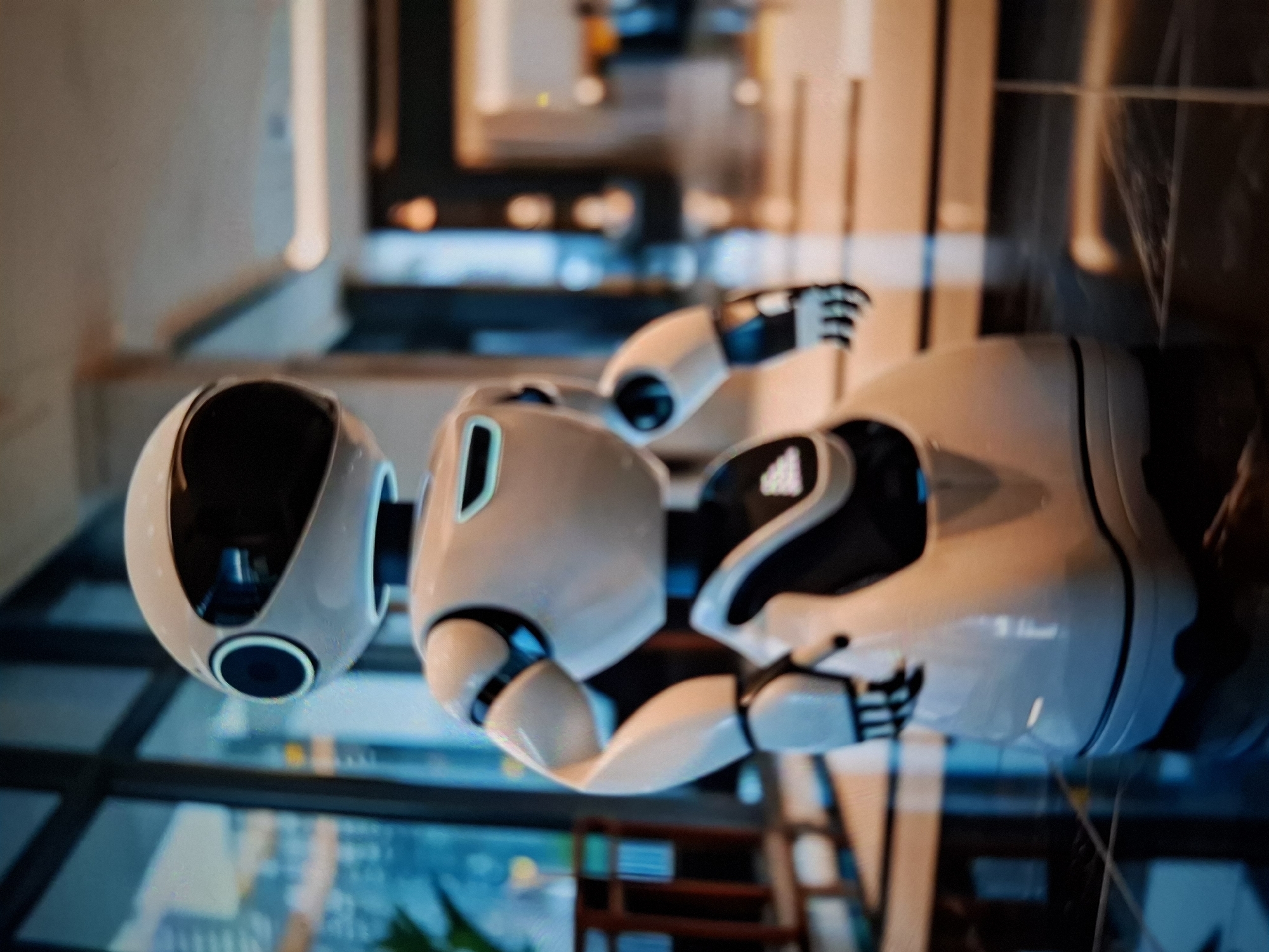 A white robot with a black visor and circular blue sensors on its head stands on a dark, reflective floor. The robot has a smooth, rounded body and articulated arms with black fingertips. It is positioned in what appears to be a modern indoor space, possibly a hotel lobby or a futuristic office, with large windows to the left showing a blurred cityscape or building interior. The background is out of focus, with soft lighting and muted colors, suggesting an urban environment in a city of unknown location. The robot is the sole prominent subject, facing forward with its head slightly tilted.