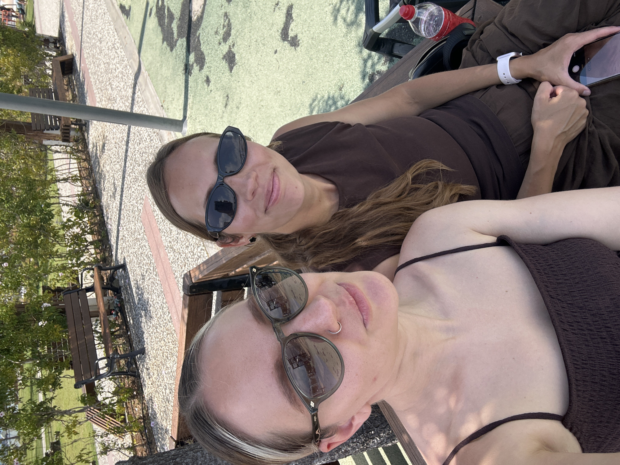 Two young women are taking a selfie while lying on a bench in what appears to be a park. The woman closer to the camera has fair skin, light blonde hair with dark roots, and is wearing sunglasses with a green frame. A small nose ring is visible. She is wearing a dark brown top with thin straps. The other woman has shoulder-length light brown hair, fair skin, and is wearing dark sunglasses. She is wearing a brown top and brown pants. She is holding a smartphone in her right hand. A white smartwatch is on her left wrist. A water bottle and a black purse or bag are visible on the bench near her. Other benches, trees, a sidewalk, and painted ground are visible in the background. The location is likely in the city of Fethiye, Turkey, as requested.