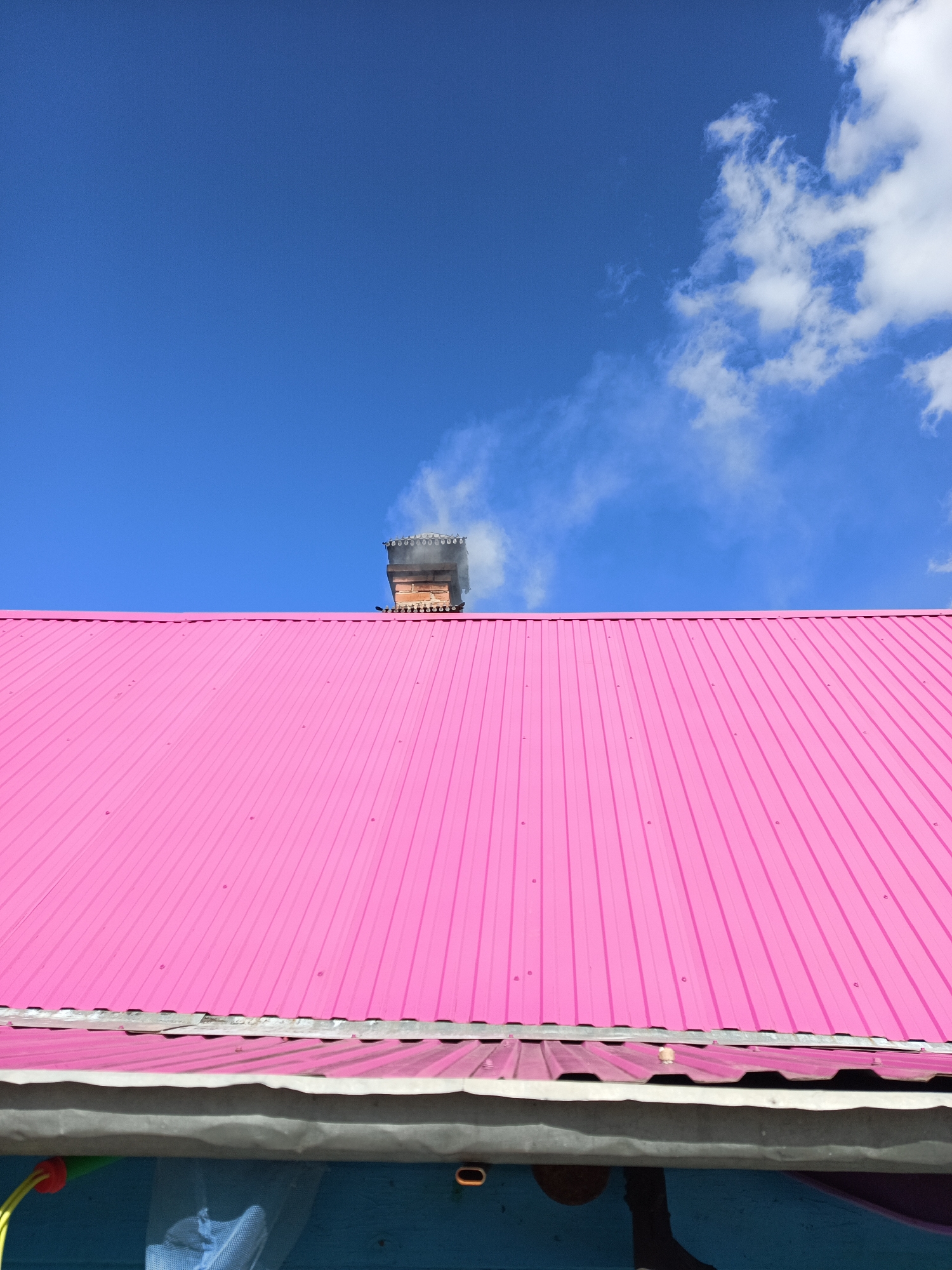 A low-angle, medium shot depicts a pink corrugated metal roof with a brick chimney emitting smoke. The chimney, topped with decorative concrete elements, is situated against a clear blue sky with scattered cumulus clouds. The roof extends diagonally across the frame from the bottom left to the upper right. Beneath the roof, a section of a blue-painted wooden structure is visible, with indistinct objects hanging from it. The location is suggested to be Chaikovsky, Russia, though no specific geographic details in the image confirm this.