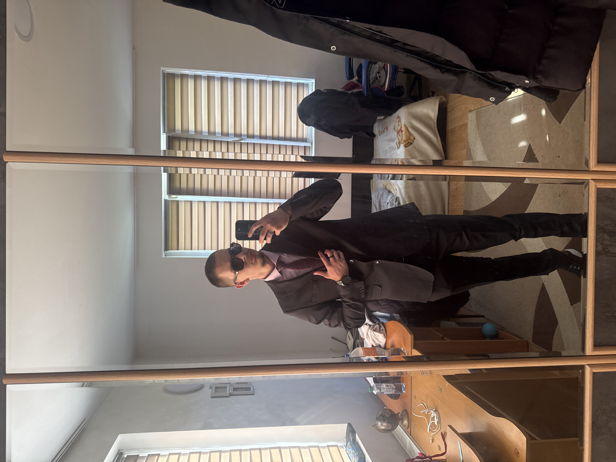 A man in a dark suit, light pink shirt, and dark sunglasses takes a selfie in a room with a desk and a mirror. The location is likely Vetren Dol, Bulgaria, based on the prompt's constraints. Blinds are visible through the window. The mirror is framed with a light-brown wood. A coat is hanging above the mirror.