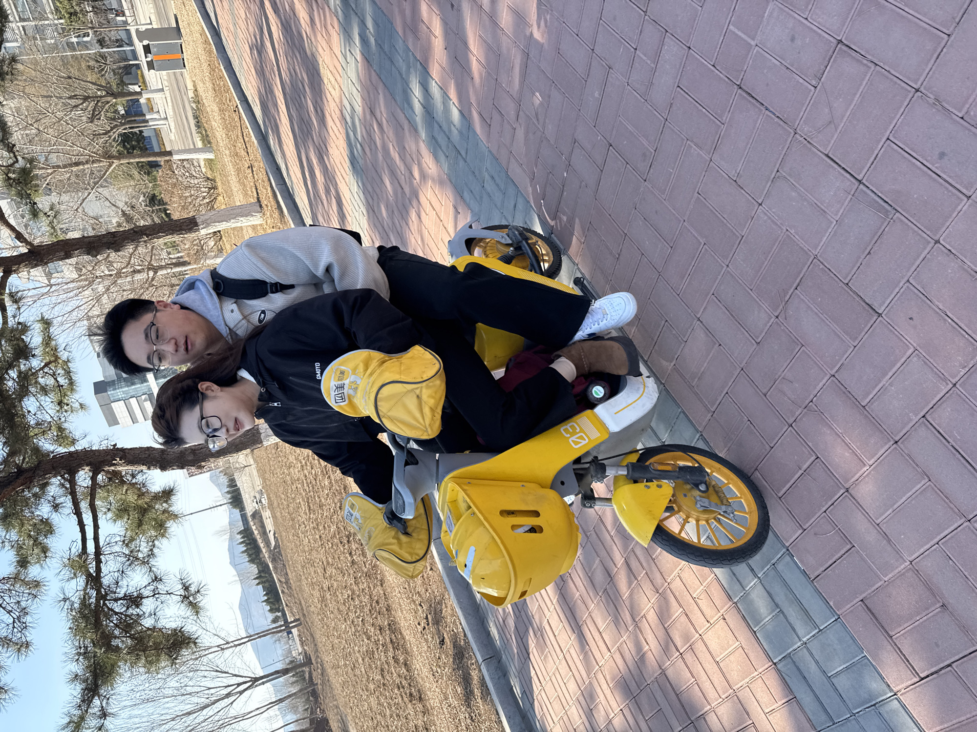 Two individuals are depicted on a yellow, child-sized tricycle on a paved brick path in Zhongtun, China. The path is bordered by grass and trees. The person in the back is wearing glasses, a light-colored hooded sweatshirt, and a backpack. The person in the front is wearing glasses and a black jacket, also seated on the tricycle. The tricycle has two yellow storage compartments. The image also shows a building in the background.